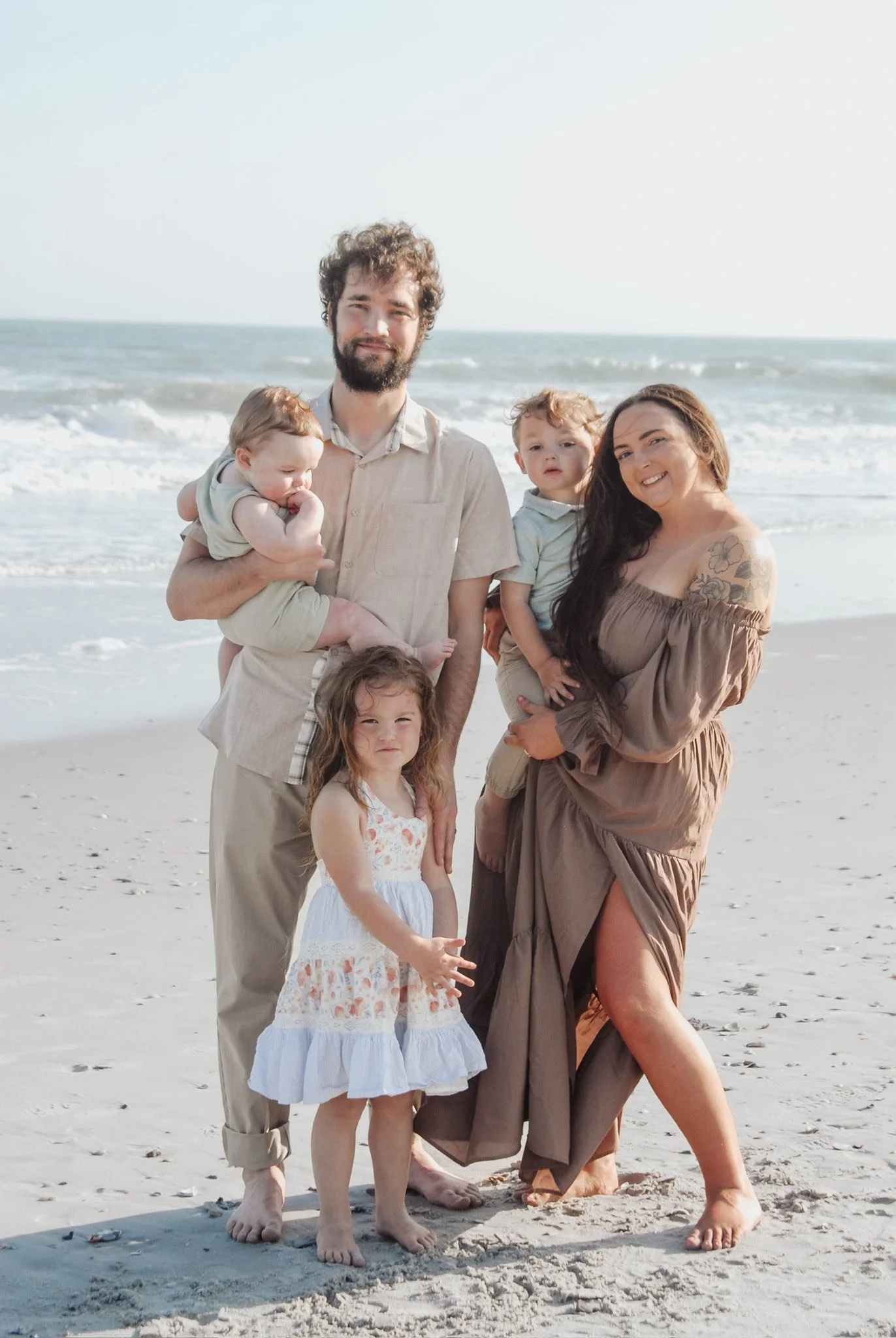 “Family beach portrait by Faith K Photography in Myrtle Beach, SC featuring parents with three young children posing on the shoreline in soft neutral tones with ocean waves in the background.”