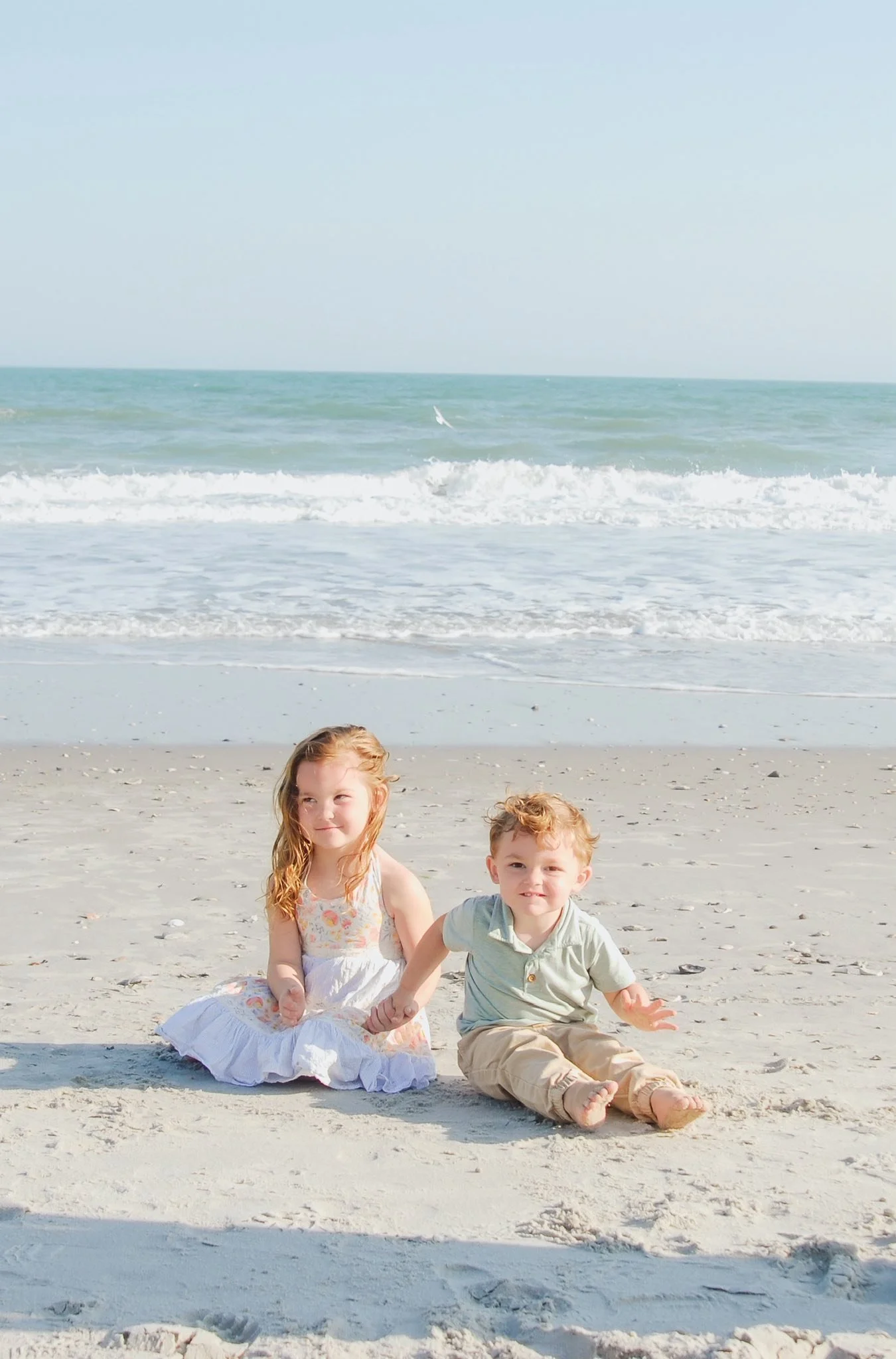“Sibling beach portrait by Faith K Photography in Myrtle Beach, SC featuring a young girl and boy sitting on the sand holding hands with ocean waves in the background.”