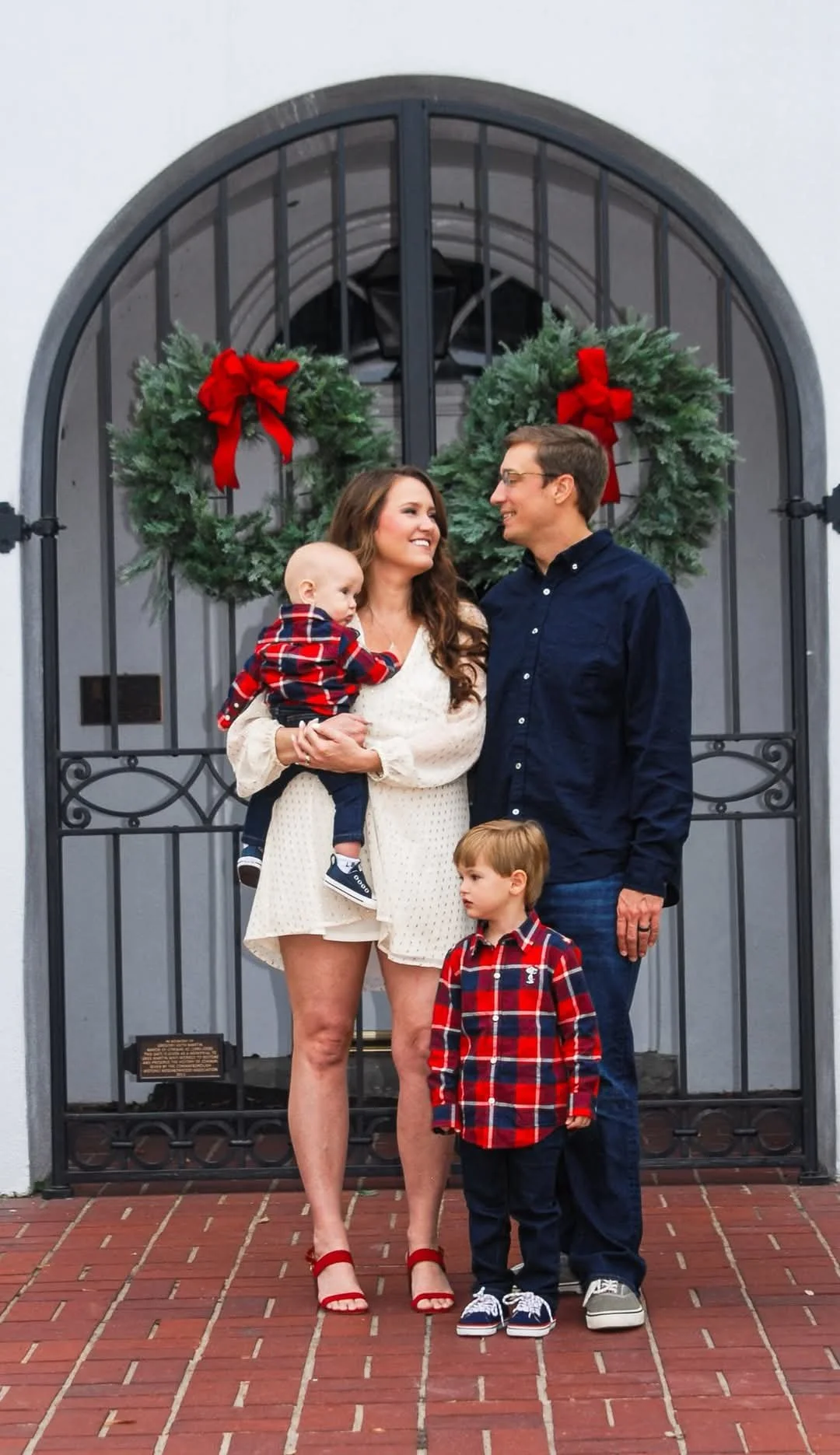 “Family Christmas portrait by Faith K Photography in Myrtle Beach, SC featuring parents with two young children in front of a festive doorway with holiday wreaths and red bows.”