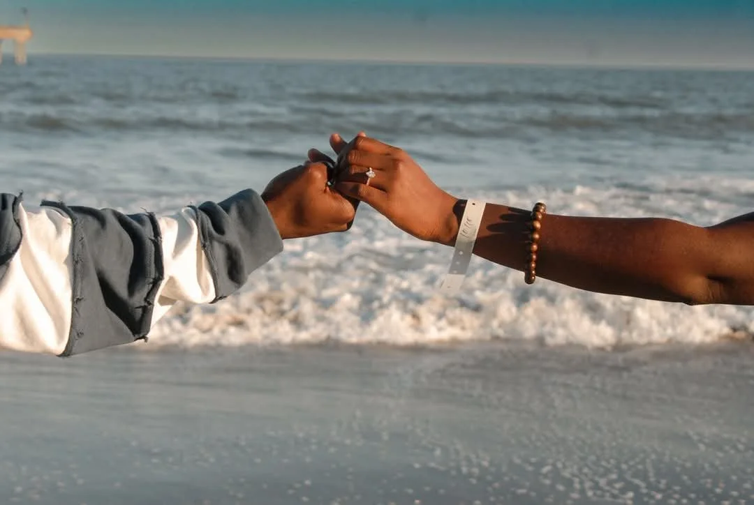 “Close-up engagement ring moment photographed by Faith K Photography in Myrtle Beach, South Carolina showing couple holding hands together on the beach with ocean waves in background.”