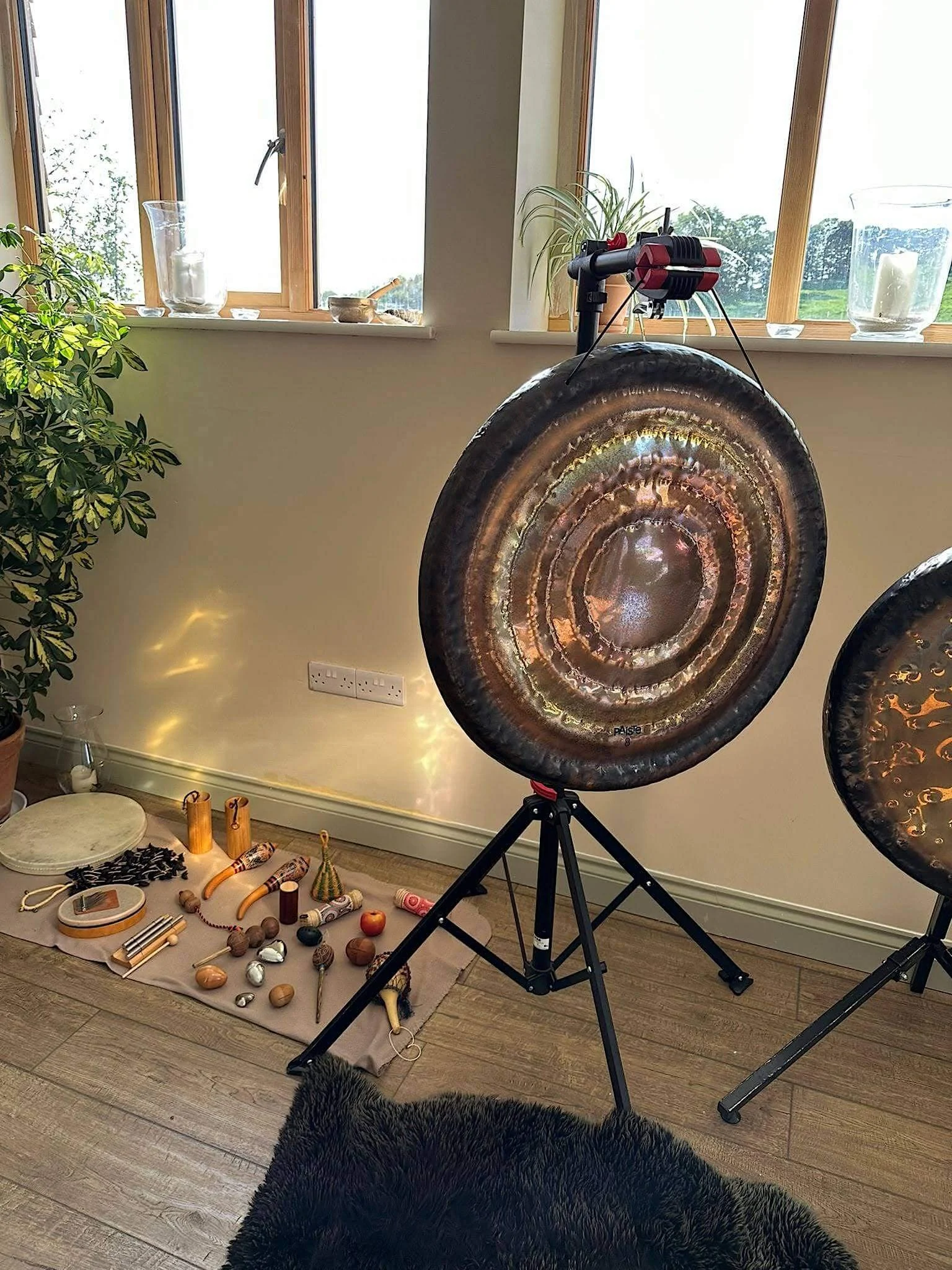 A room with two large gong instruments on stands, a window with plants and candle holders, and a collection of small percussion instruments and objects on a cloth on the floor.