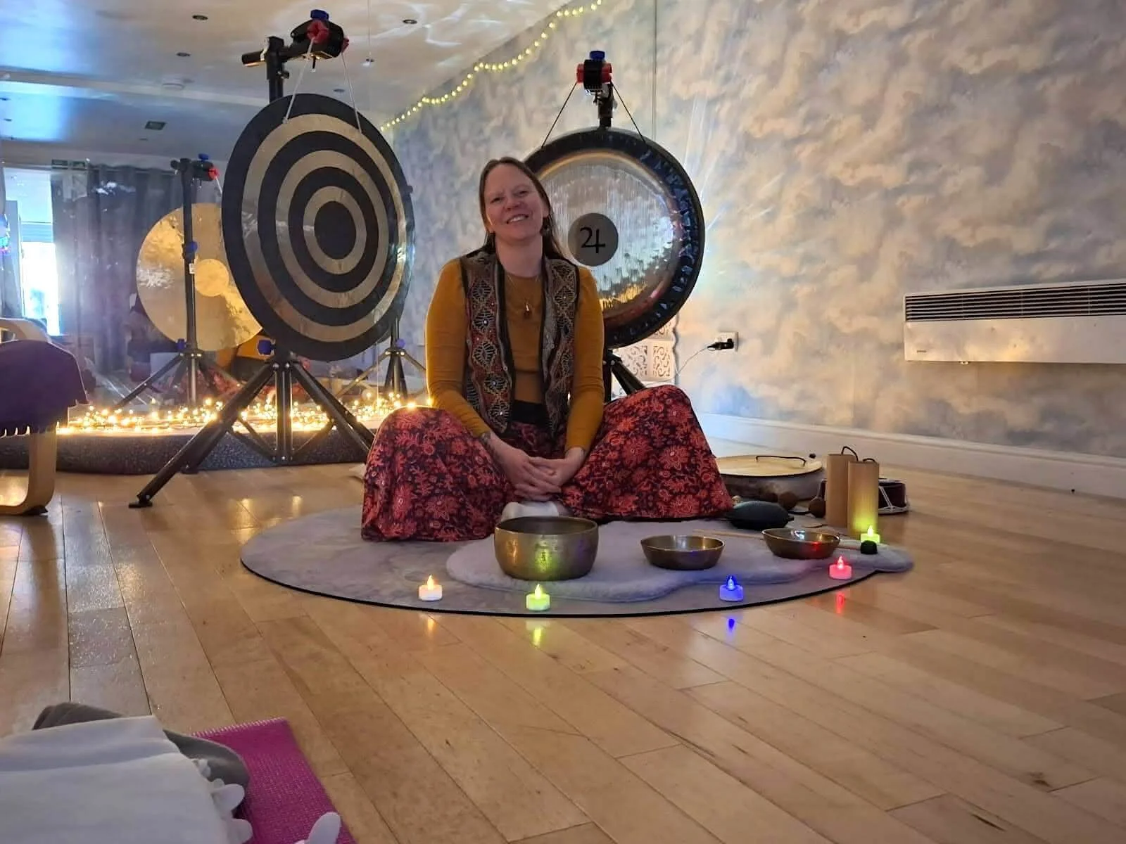 Woman sitting cross-legged on a round mat with singing bowls and candles, smiling at the camera, with large crystals and a gong in the background, in a room with wooden flooring and a blue cloud-like patterned wall.