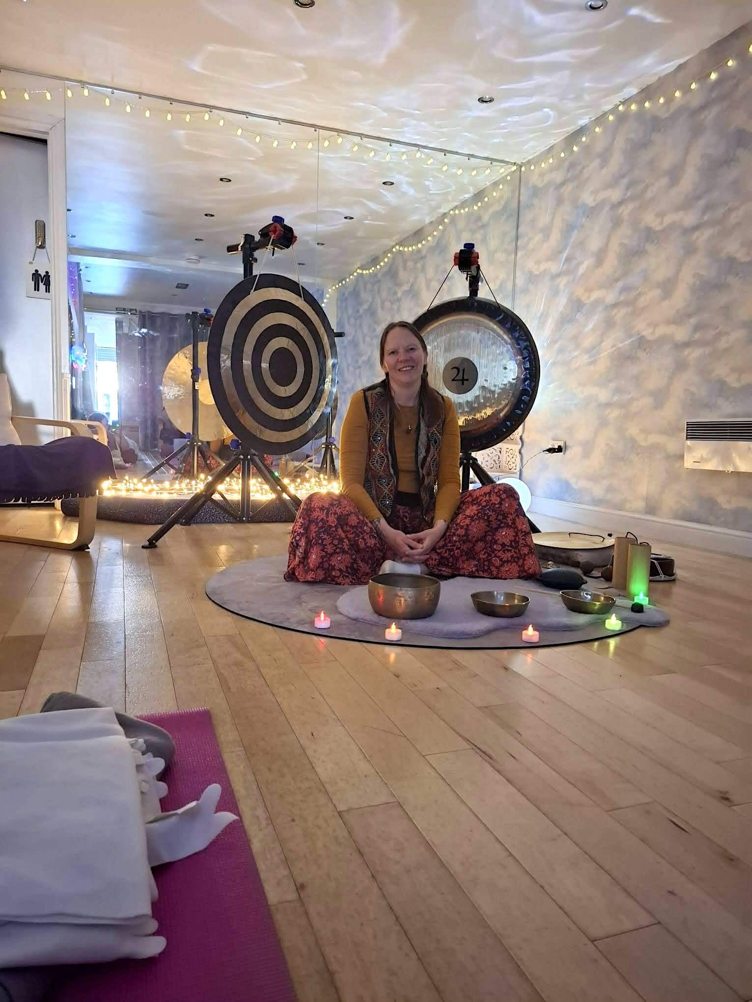 Woman sitting cross-legged on a round rug in a decorated indoor space with goal tarts, singing bowls, and candles, surrounded by string lights and two large targets in the background.