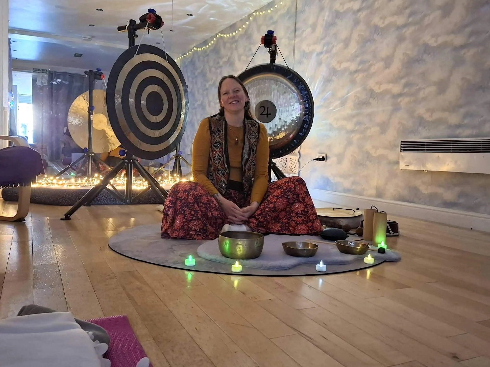 A woman sitting cross-legged on a round rug surrounded by singing bowls and candles, smiling in front of large gong drums with bullseye targets, illuminated by warm and soft lighting in a cozy indoor setting.