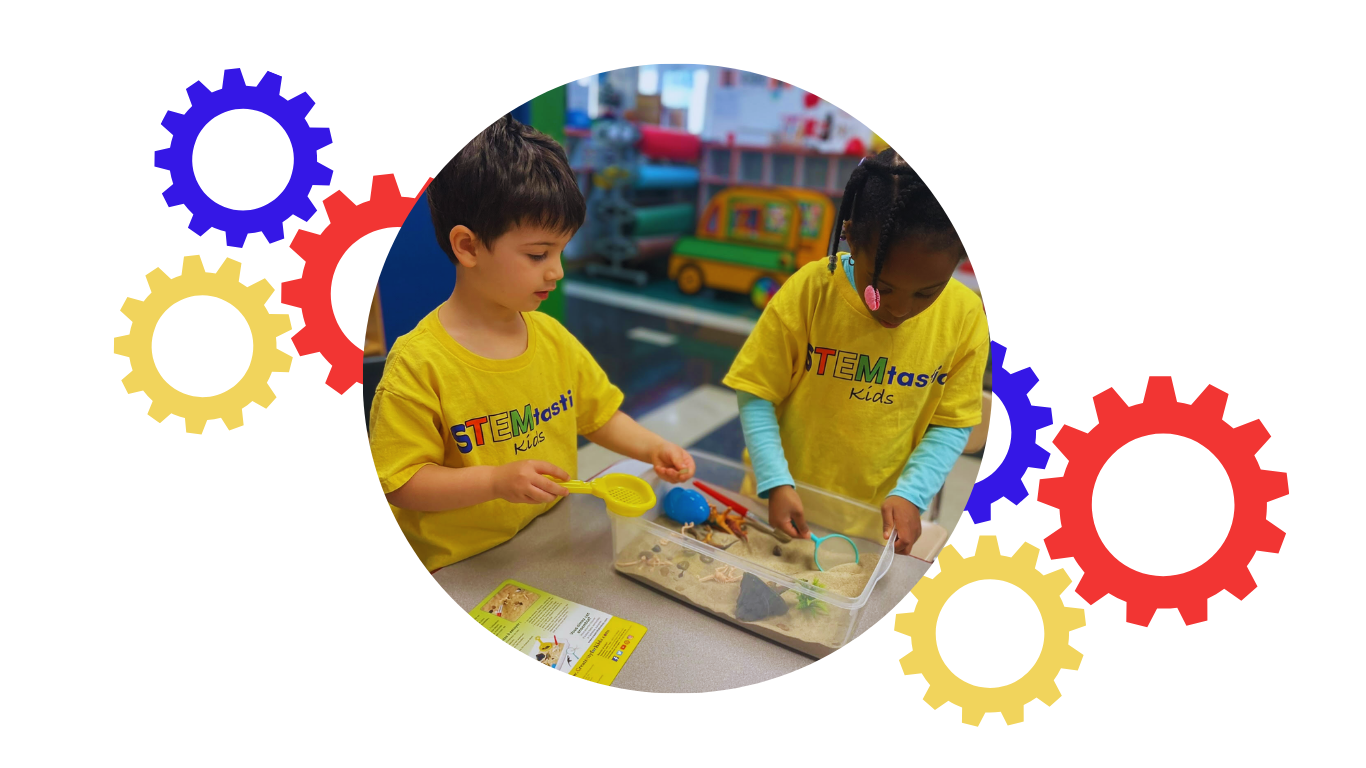 Two children wearing yellow STEM T-shirts exploring a sandbox with various objects inside at a STEM activity center.