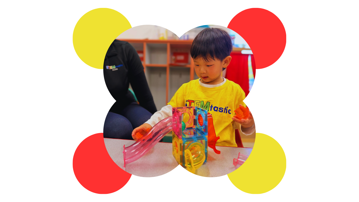 A young boy in a yellow shirt playing with colorful marble run toys on a table indoors, with a blurred person nearby and a background of shelves and vibrant colors.