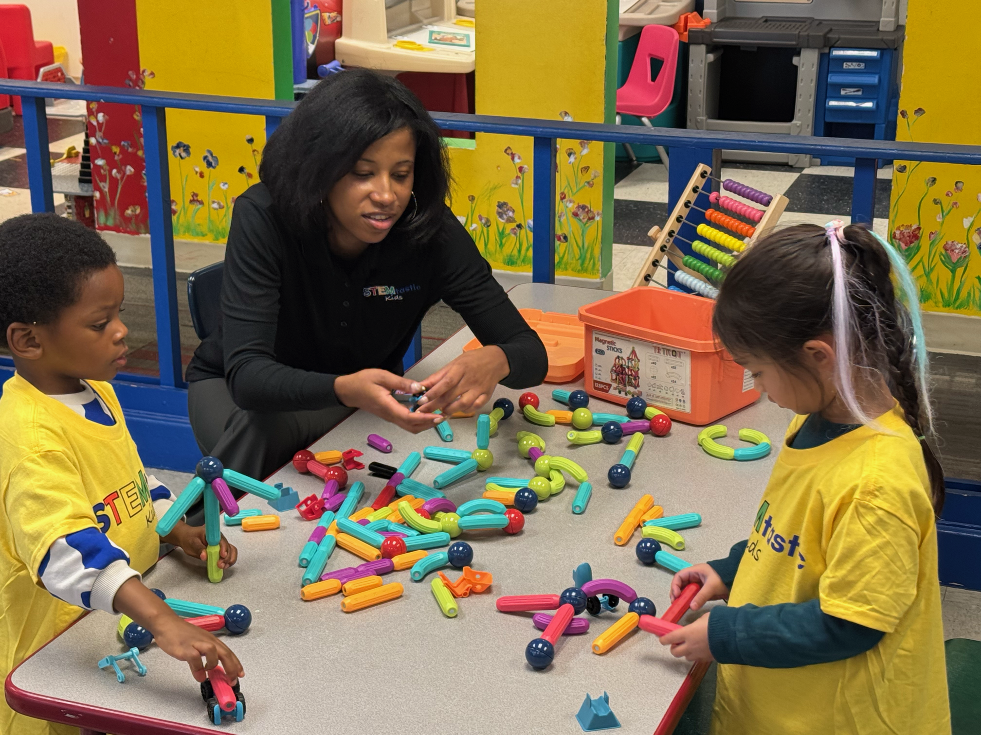 A group of children and an adult sitting at a table playing with colorful building toys in a classroom decorated with bright yellow walls and painted flowers.