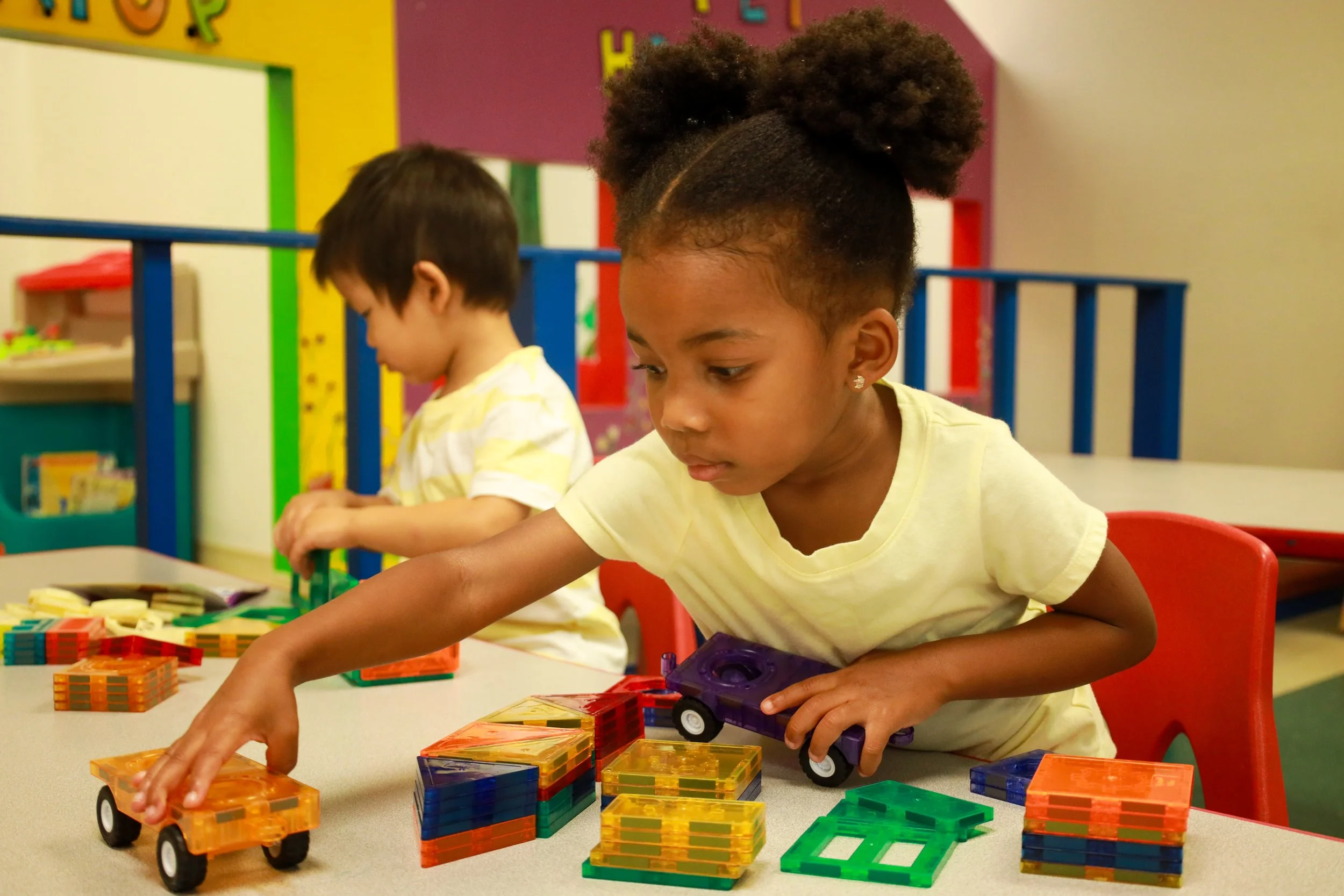 Two young children playing with colorful building blocks and toy trucks at a table in a classroom.