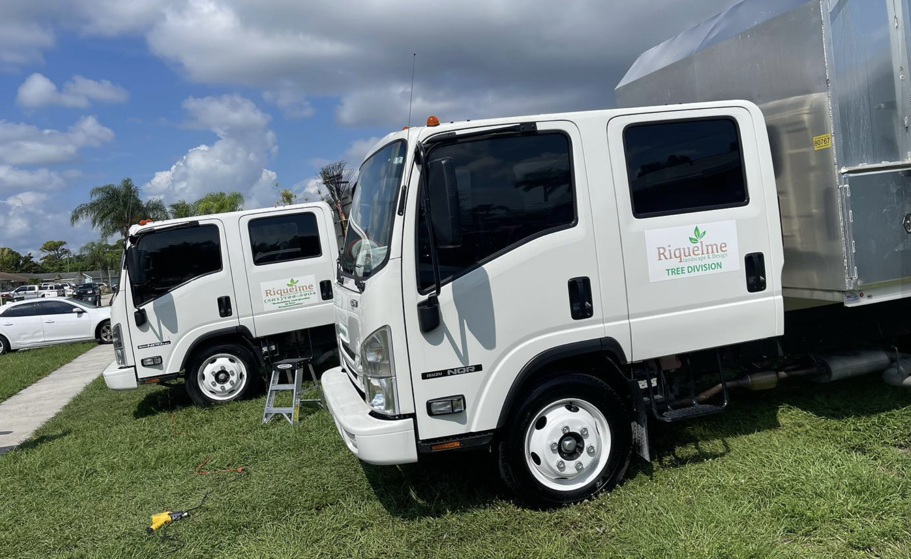 Two white trucks with 'Riquelme Landscape & Design Tree Division' logos parked on a grassy area with a cloudy sky and a parking lot in the background.