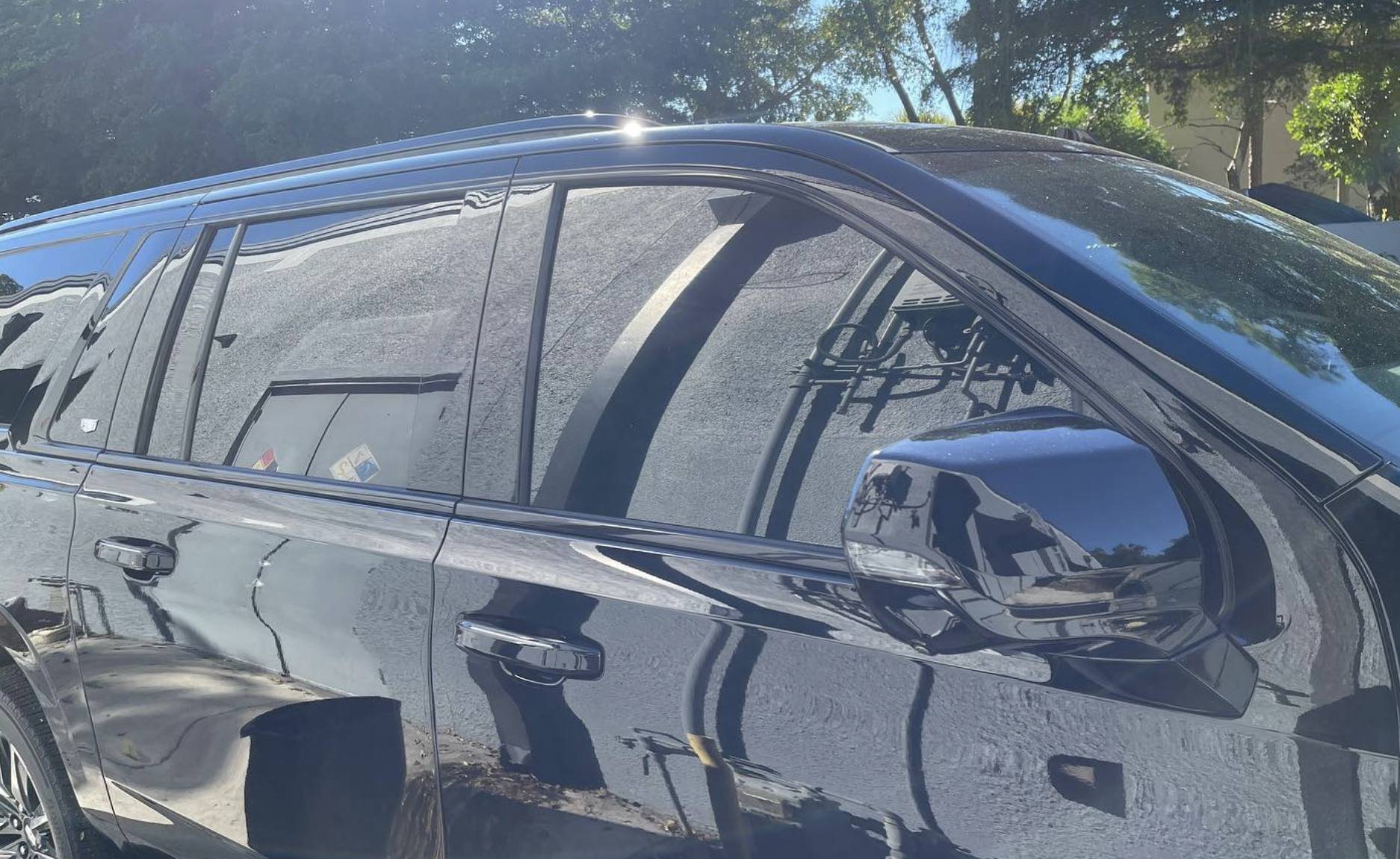 Close-up of a shiny black SUV, showing reflections of trees, sky, and buildings on its surface and windows, parked outdoors on a sunny day.