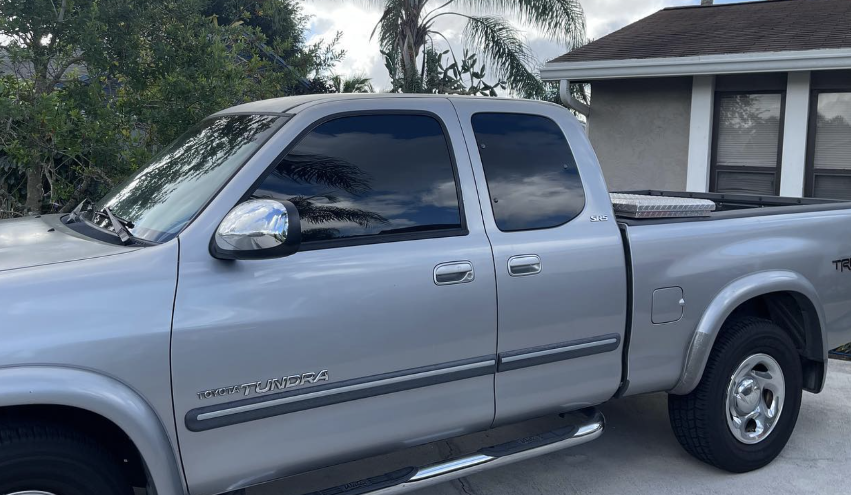 Silver Toyota Tundra pickup truck parked outside near a house with a gray exterior and dark window blinds, with trees and bushes in the background.