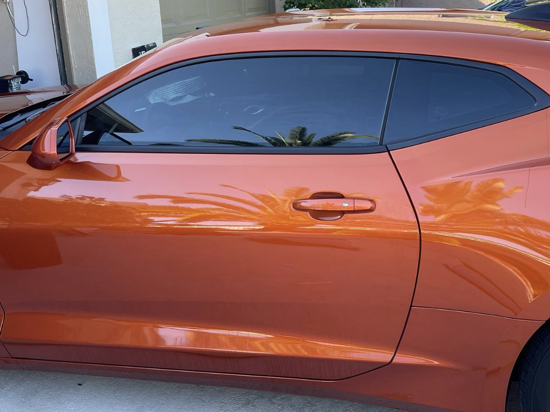 Orange sports car parked near a garage door, with reflections of palm trees on its windows and body.