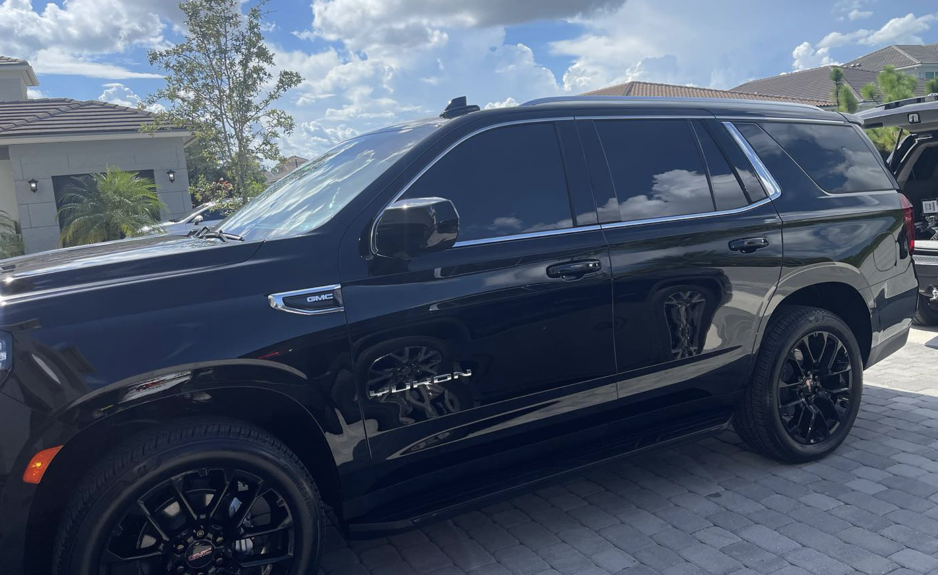 Black GMC Yukon SUV parked on a brick driveway in front of a house with a blue sky and clouds reflected on its windows.