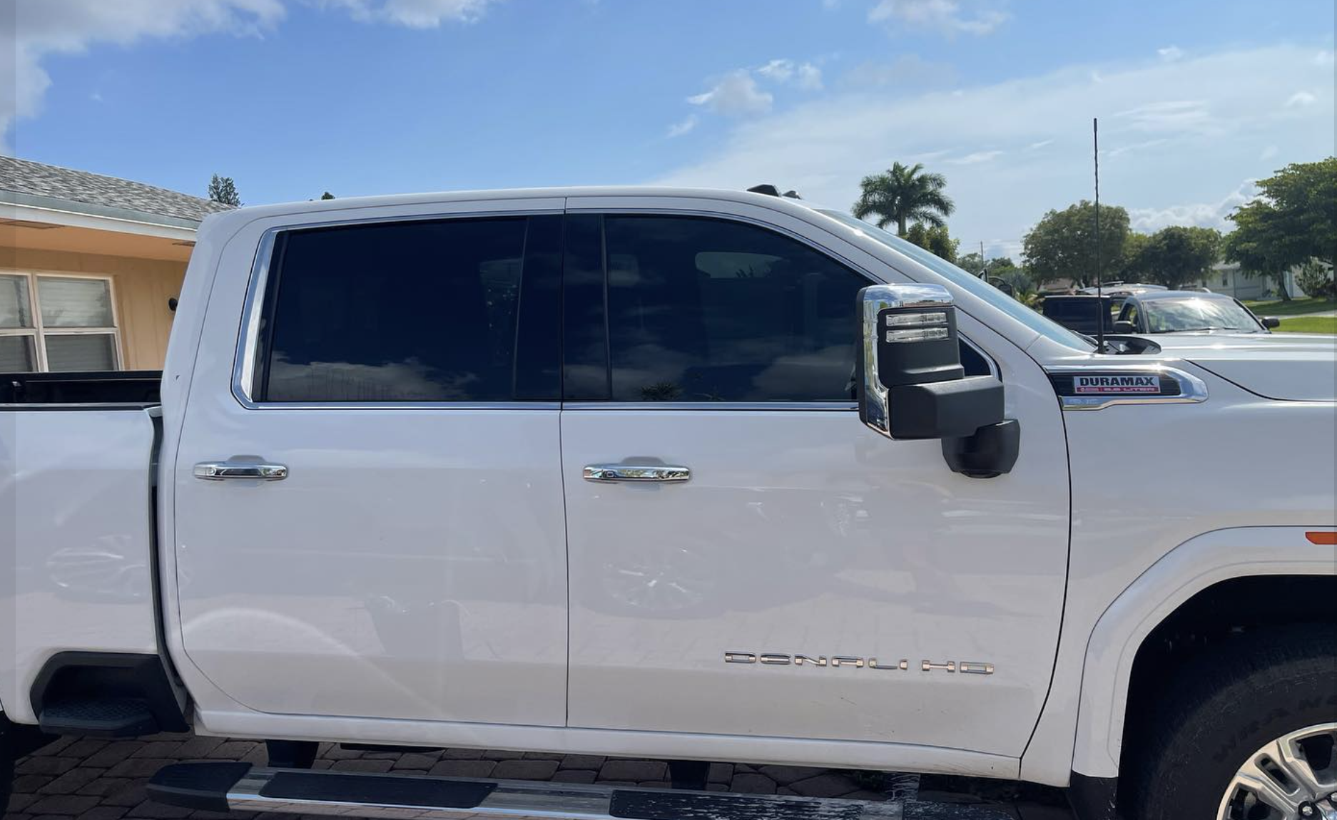 A white GMC Denali pickup truck parked outdoors on a sunny day with a house and trees in the background.