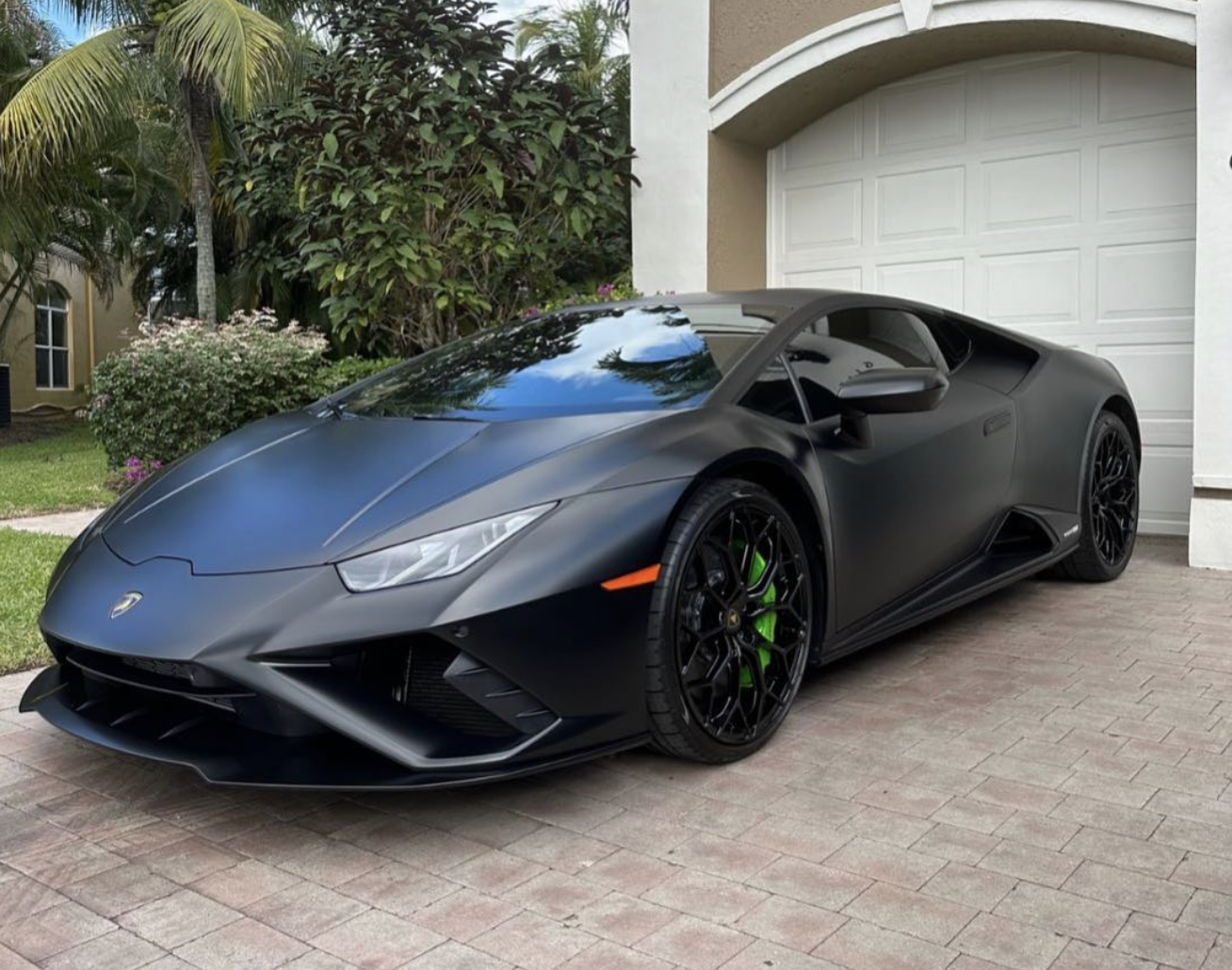 Black Lamborghini sports car parked on a brick driveway in front of a white garage door, with green foliage and flowering plants in the background.