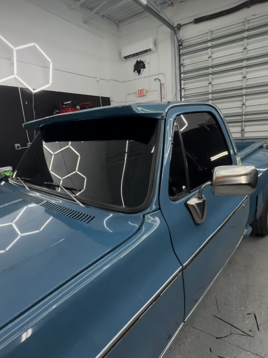 A vintage blue pickup truck parked inside a garage with hexagon-shaped LED wall lights reflecting off the windshield and the body of the vehicle.