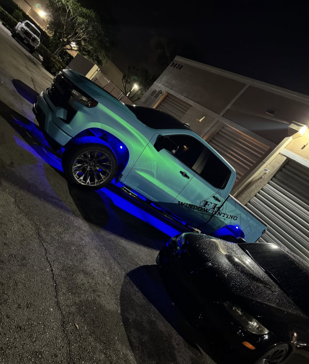 A pickup truck with custom lighting reflects green and blue underglow lights, parked in an outdoor lot at night. The truck has a decal that reads 'Window Tinting' on its side. Nearby are other cars and a building with a garage door.