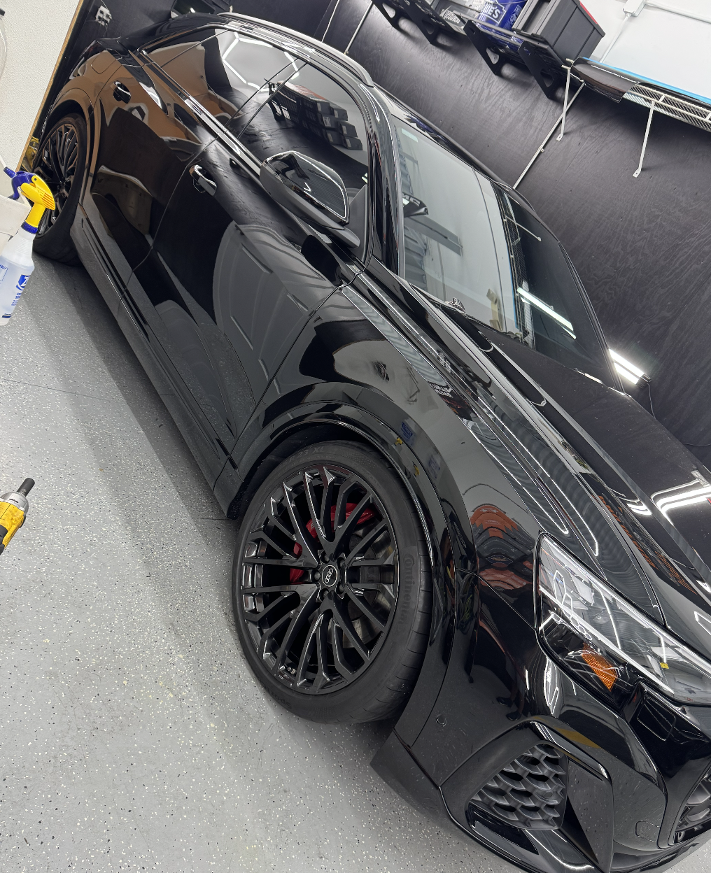 A black sports car parked indoors on a gray speckled floor, with cleaning supplies nearby, reflecting the ceiling lights and surroundings on its shiny surface.