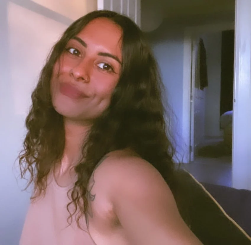 A woman with long, curly hair smiling at the camera in a room with soft lighting.