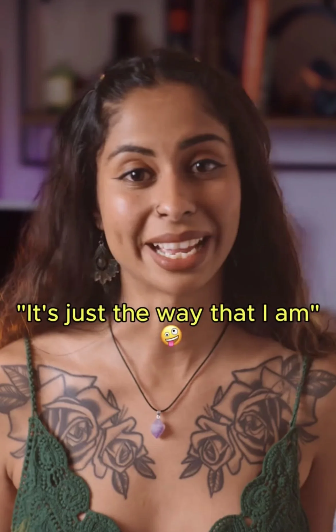 A woman with long wavy hair, tattoos of roses on her chest, wearing a green lace top and a purple gemstone necklace, speaking with a smile. Subtitles say, 'It's just the way that I am' with a playful tongue-out emoji.