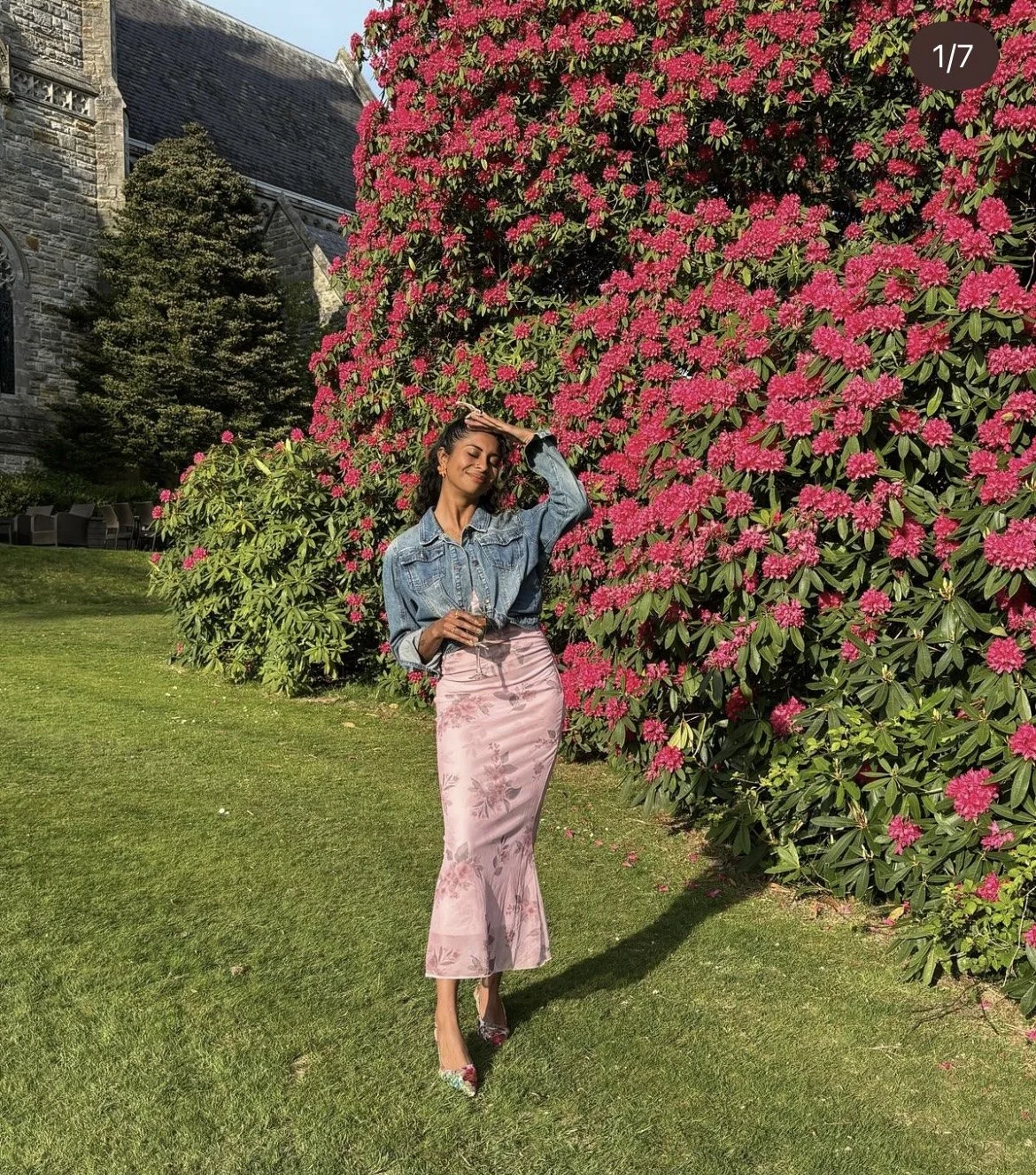 A woman in a denim jacket and pink floral skirt stands on a grassy area, smiling with her hand on her forehead, next to a large blooming pink rhododendron bush outside a stone building.