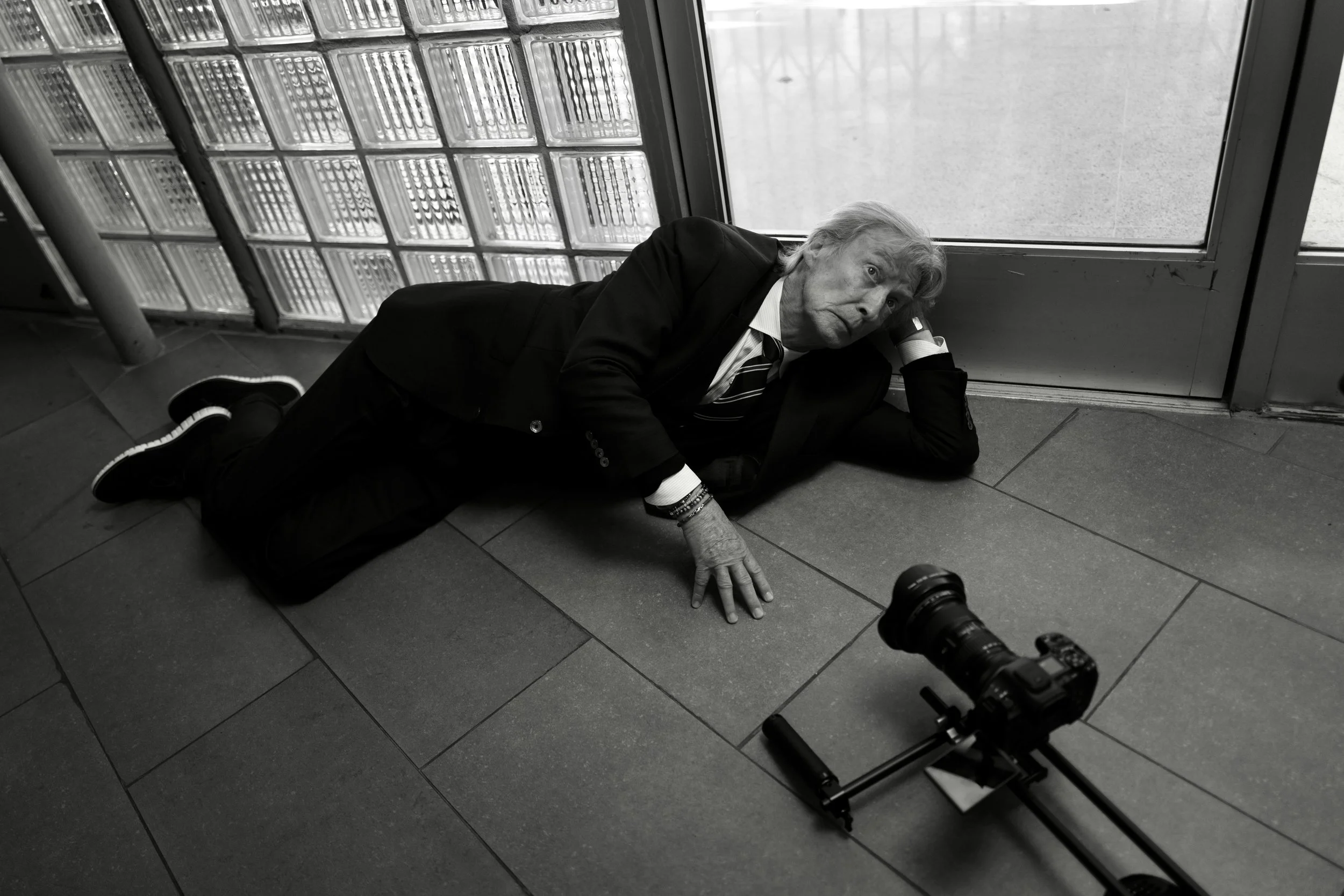 An elderly man in a suit lying on the floor with a camera mounted on a stabilizer nearby, indoors near a glass brick wall and door, looking up with a distressed expression.
