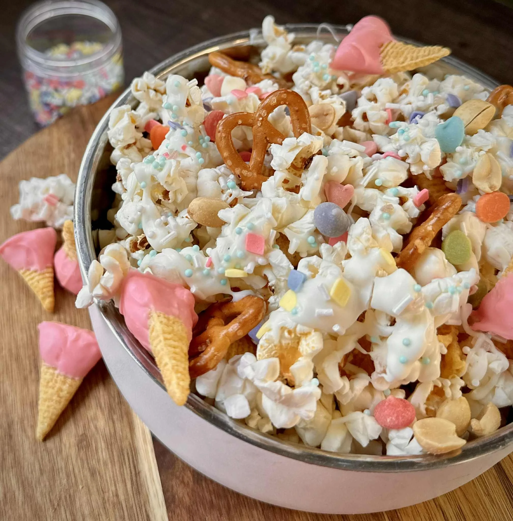 A bowl of popcorn decorated with colorful sprinkles, candy shapes, pretzels, and pink ice cream cone-shaped toppings. Some popcorn pieces and decorations are scattered on a wooden surface outside the bowl.
