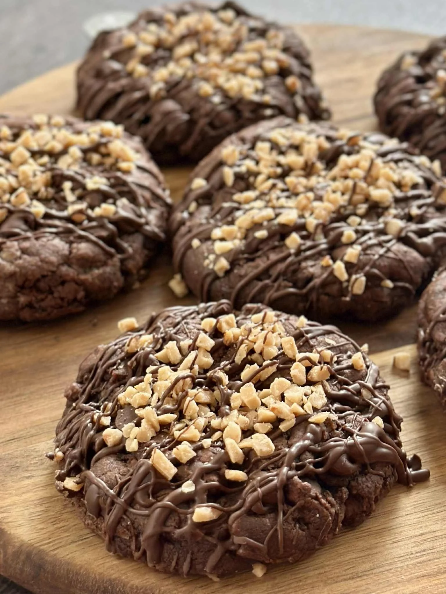 Close-up view of chocolate heath bar cookies topped with toffee pieces and drizzled with chocolate, placed on a wooden surface.