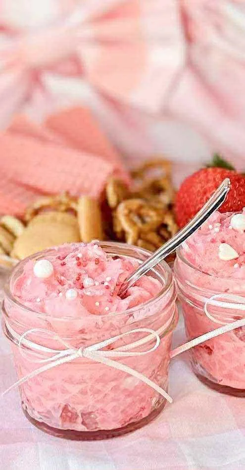 Two small jars of pink strawberry-flavored whipped dessert garnished with white edible pearls and a silver spoon, with strawberries and cookies in the background.