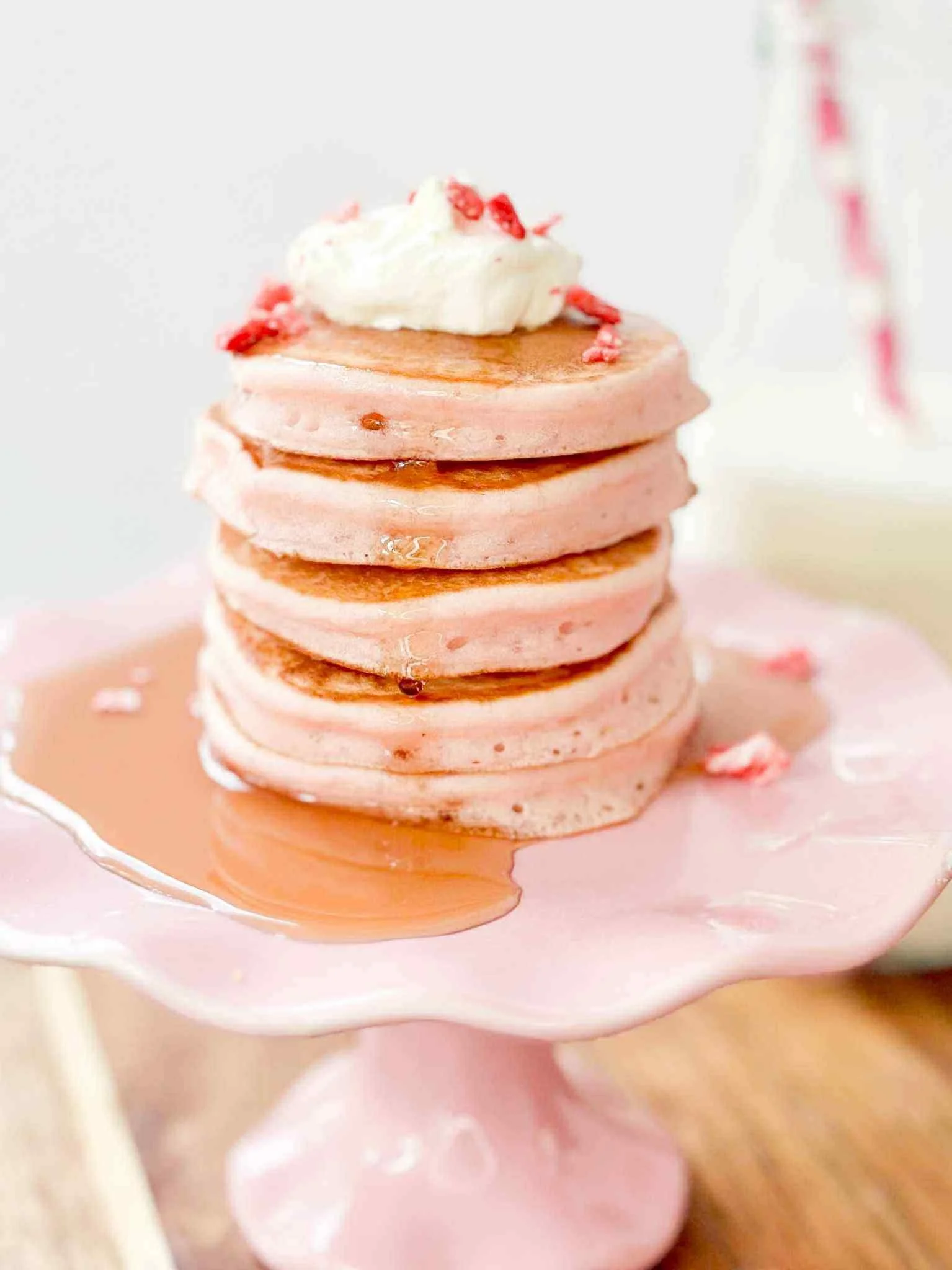 Stack of pink pancakes topped with whipped cream and small red toppings, served on a pink cake stand.