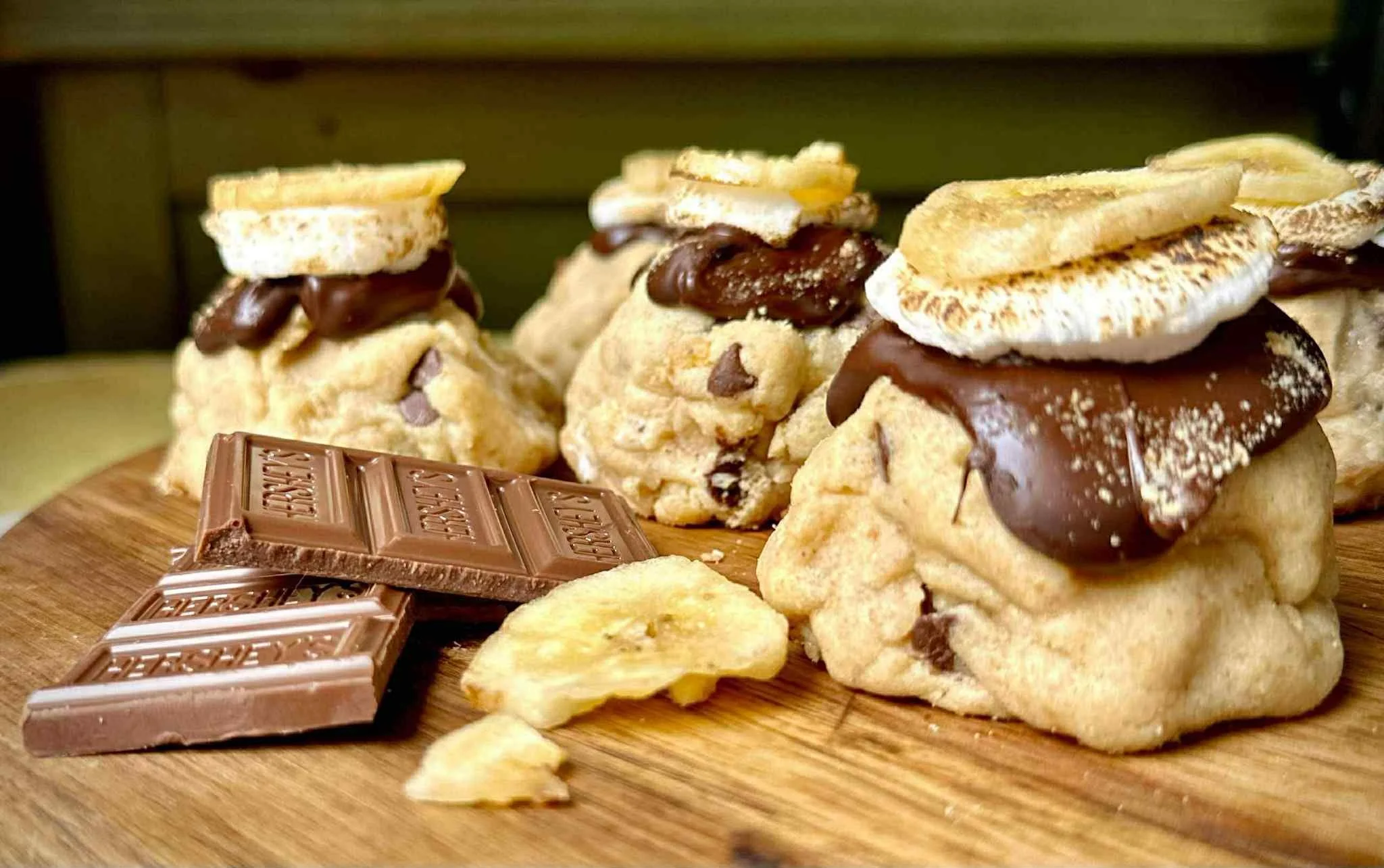 Four s'mores made with cookies, chocolate, marshmallow, and a banana slice on a wooden board, with broken chocolate pieces and banana chips nearby.