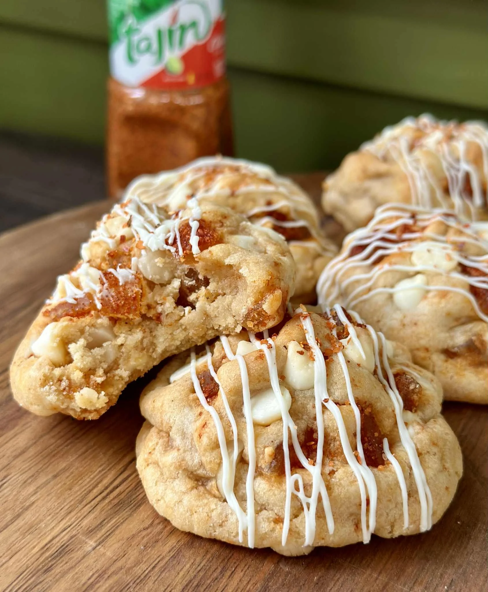 Close-up of cinnamon rolls topped with white icing and drizzled with caramel, placed on a wooden tray with a bottle of Tajín seasoning in the background.