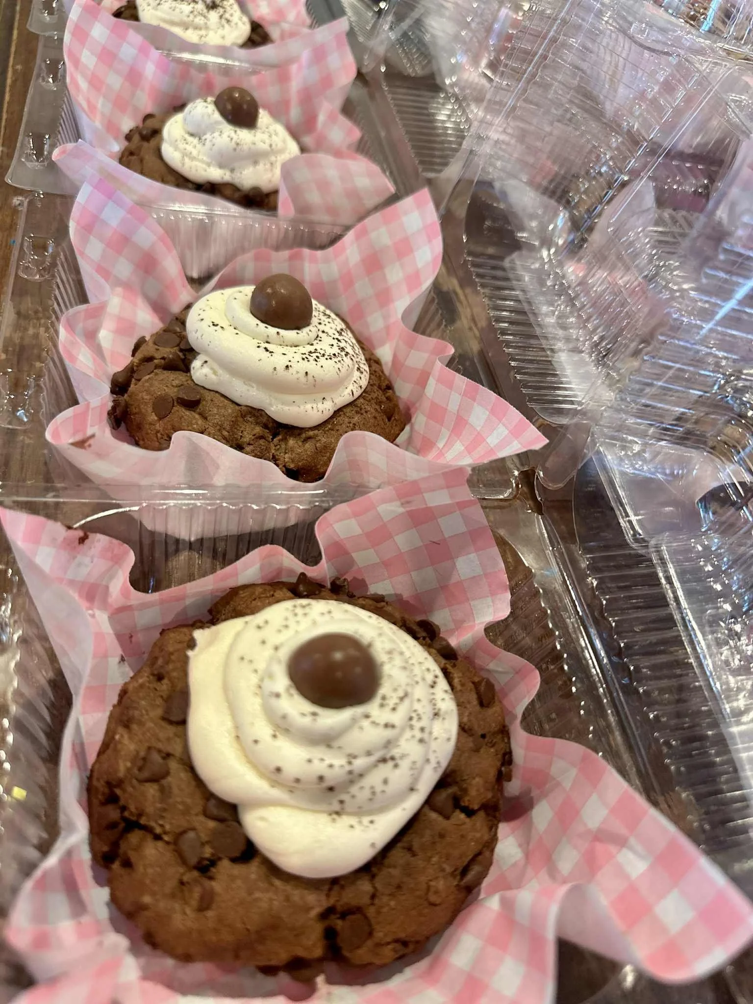 Three chocolate cookies with white whipped cream and a chocolate round on top, placed in pink checkered paper cups inside a clear plastic container.
