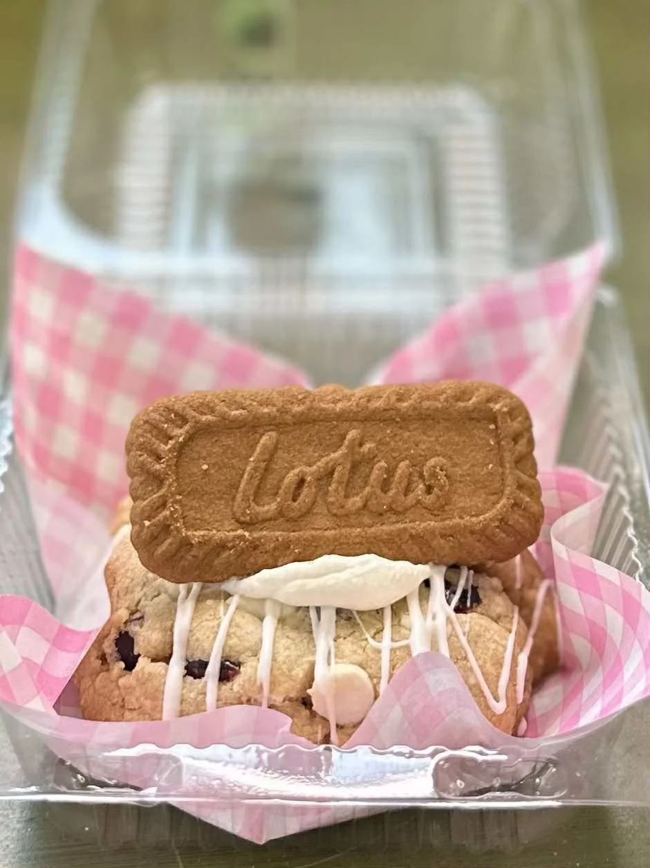 A cookie butter cranberry cookie with white icing filling, topped with a Lotus biscuit, inside a clear plastic container lined with pink checkered paper.
