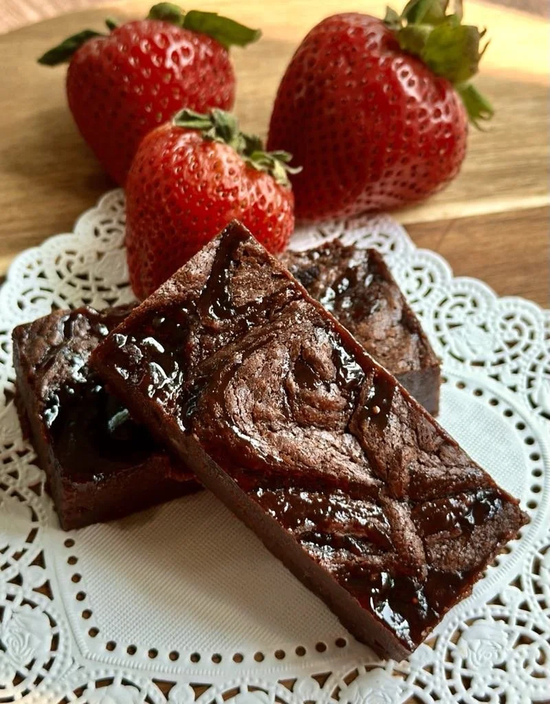 Three fresh strawberries placed on a white decorative lace doily with two pieces of chocolate brownie cake on a wooden surface.