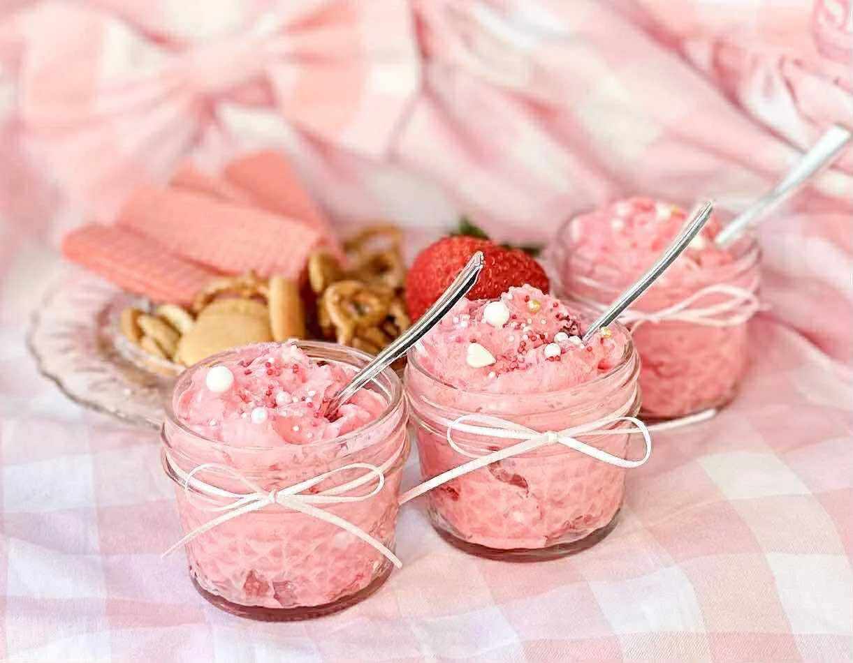 Three small glass jars filled with pink strawberry mousse, decorated with sprinkles and heart-shaped candies, each with a silver spoon. In the background, there are cookies, a strawberry, and pink wafers, all on a pink checkered tablecloth with pink 