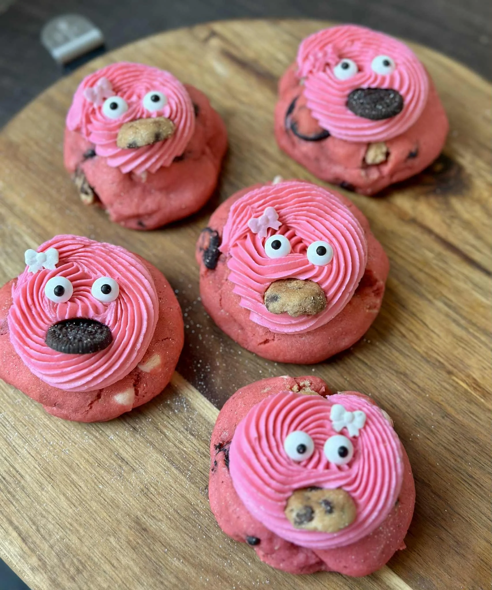 Five pink cookies decorated with pink frosting, chocolate chips, small candy eyes, and tiny white bows, arranged on a round wooden serving board.