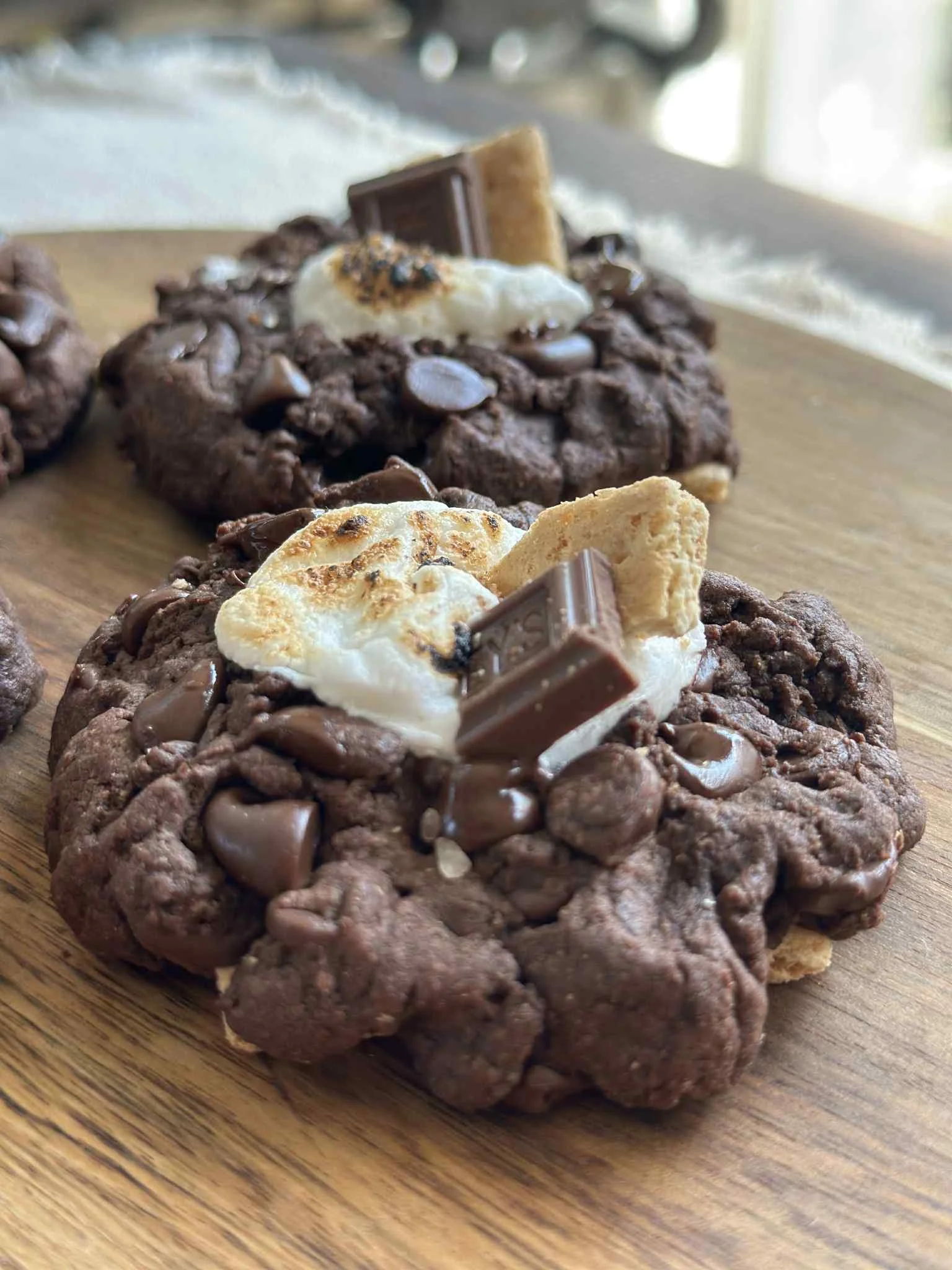 Close-up of chocolate cookies topped with marshmallows, chocolate chips, Kit Kat pieces, and graham cracker crumbs on a wooden surface.