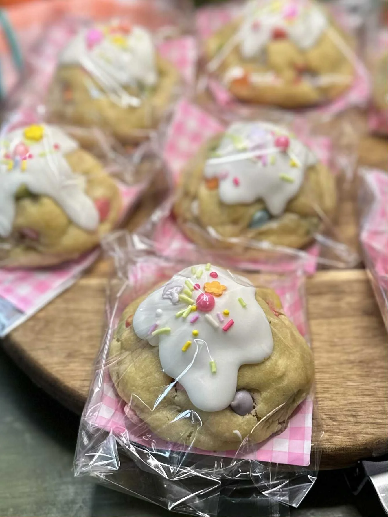 A decorated cookie with white icing and colorful sprinkles, packaged in clear plastic, placed on a pink-checked paper liner.