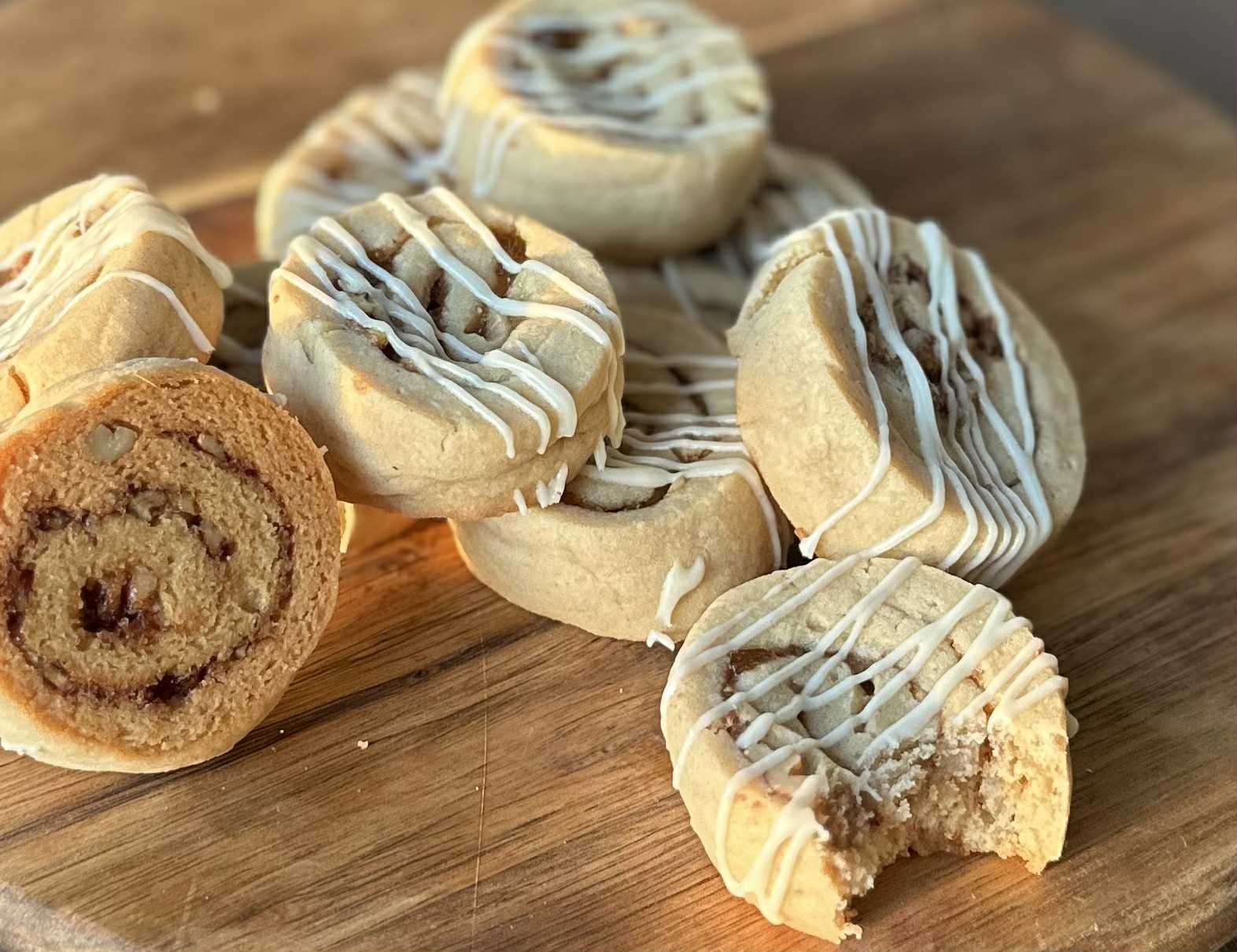Close-up of several rolled cookies with white icing drizzled on top, placed on a wooden surface, with some broken showing a chocolate chip filling.