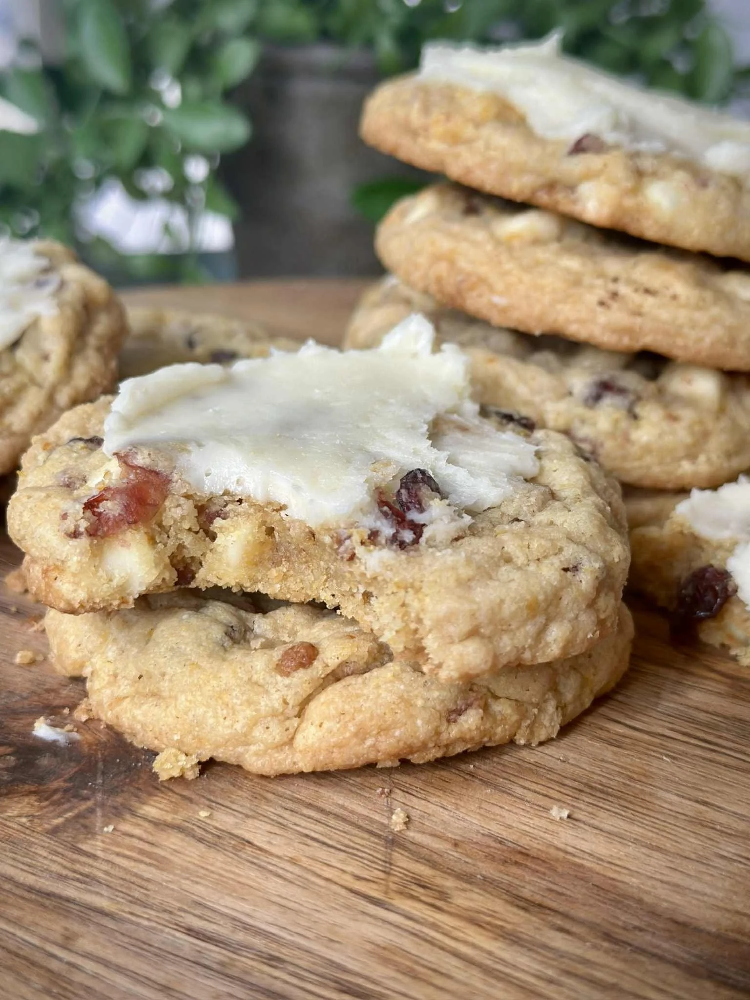 Close-up of three chocolate chip cookies with one topped with white icing, placed on a wooden surface with a leafy background.
