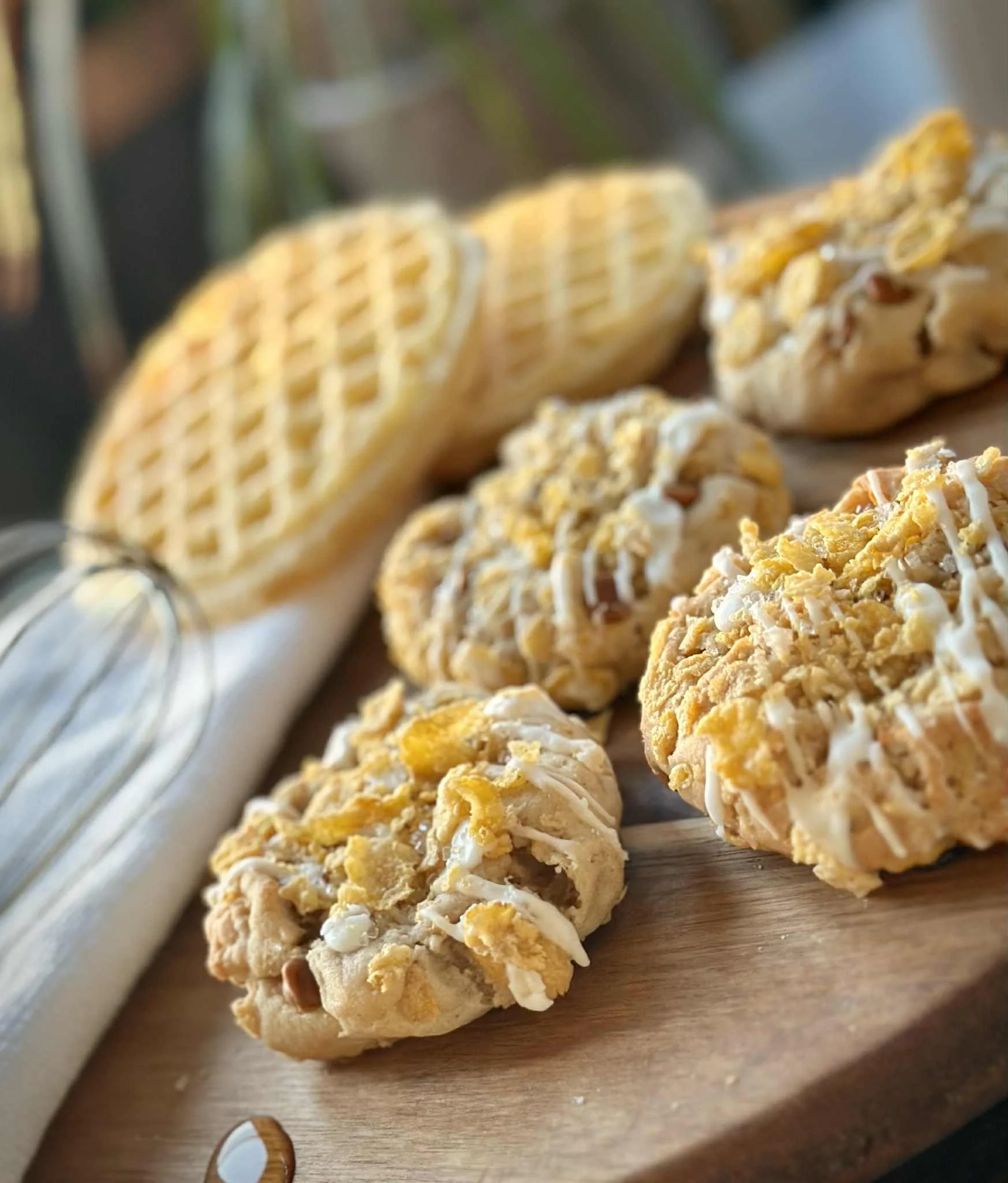 Close-up of three cookie balls topped with crumbled cookies and drizzled white icing, placed on a wooden board with waffles and a wire whisk in the background.