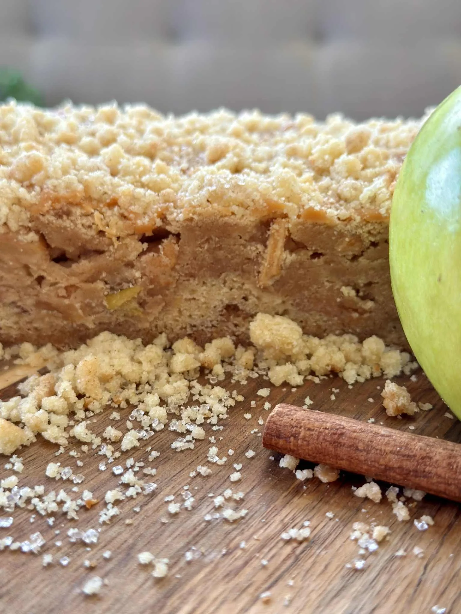 Close-up of a crumbly apple crumble dessert with a green apple and a cinnamon stick on a wooden surface.