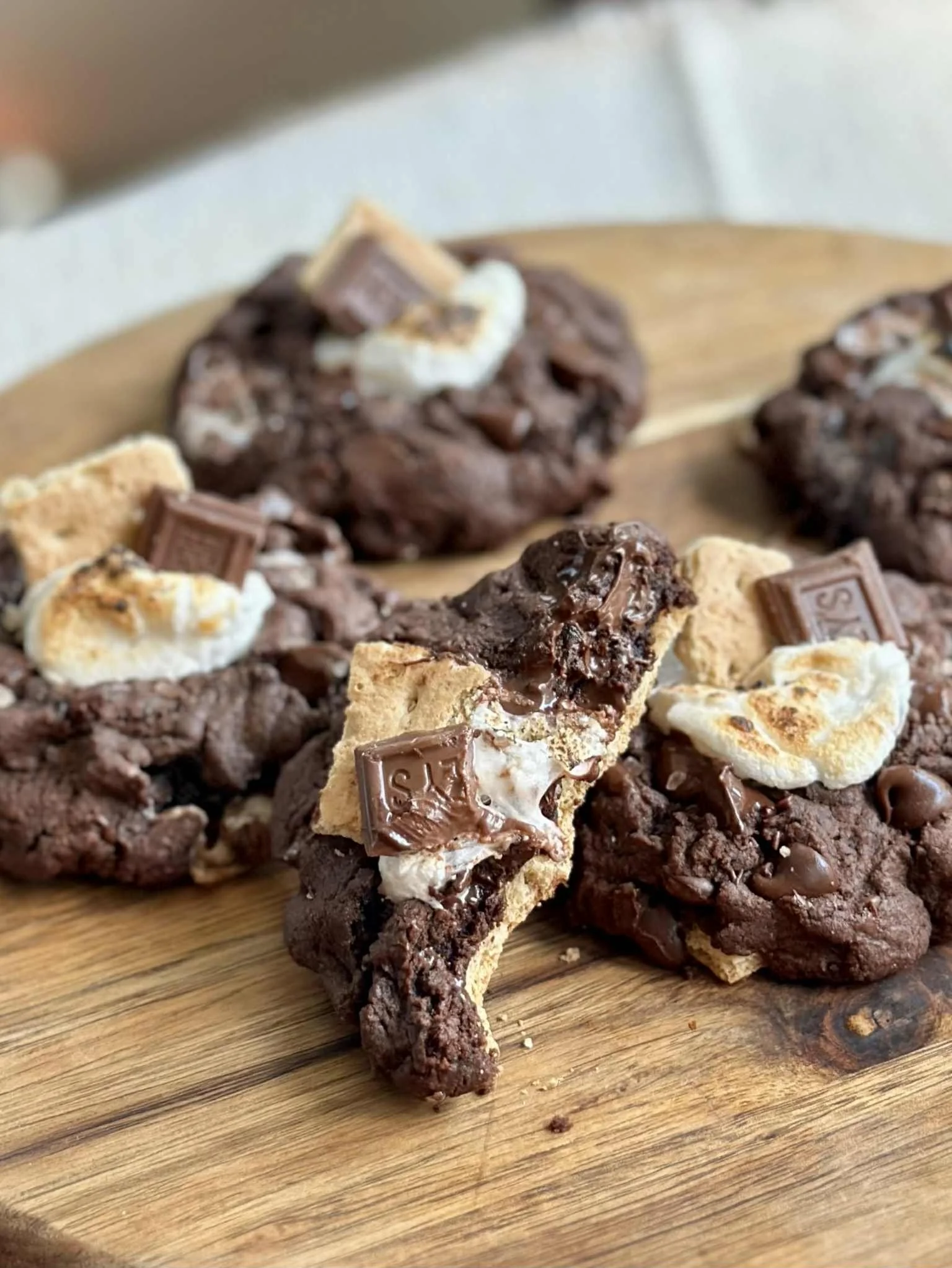 Chocolate cookies with marshmallows, graham crackers, and pieces of Snickers candy bar on a wooden cutting board.