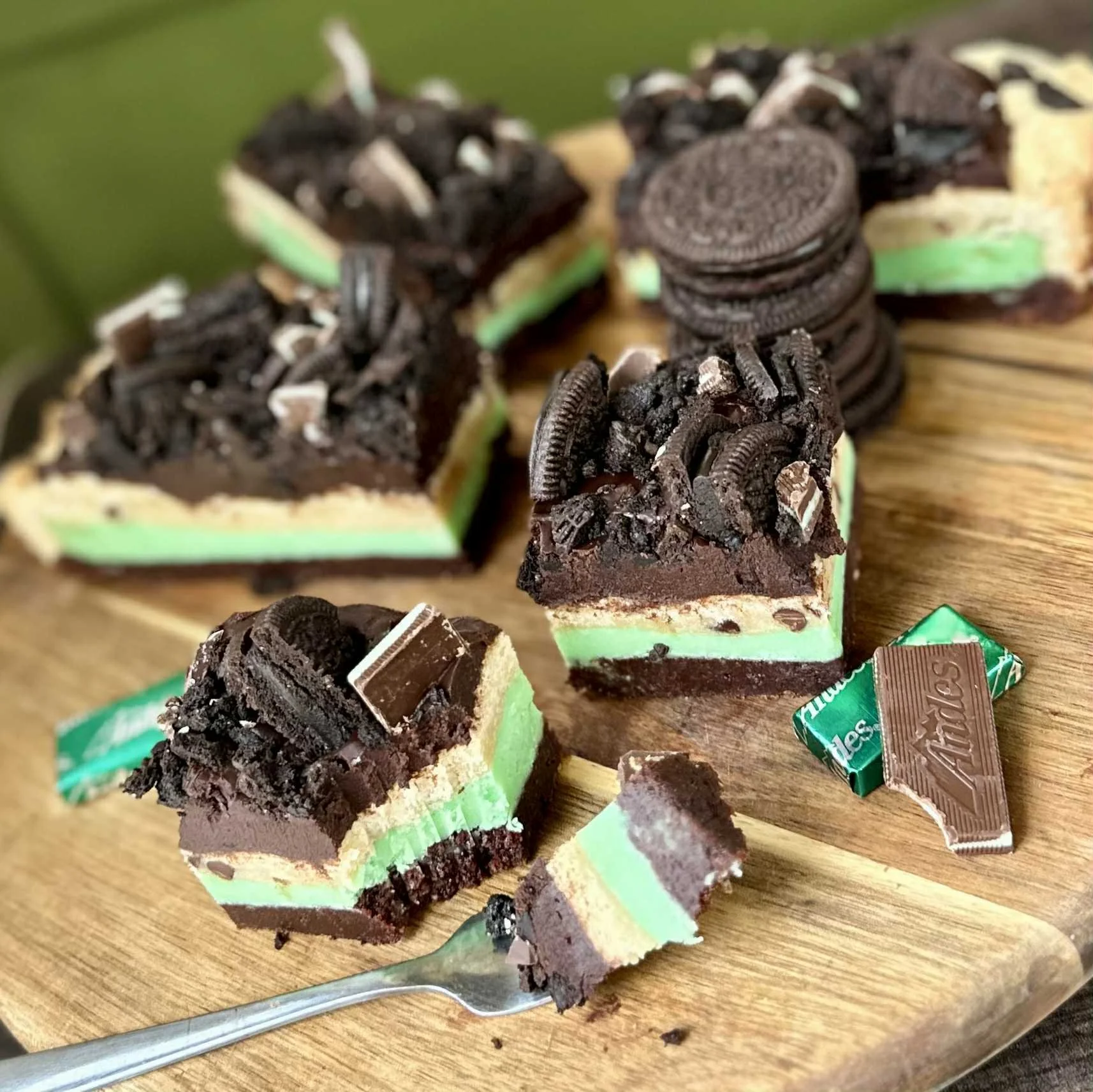 Slices of Oreo cheesecake with green and white layers, topped with crushed Oreo cookies, on a wooden cutting board, with a fork and some crumbled chocolate pieces.