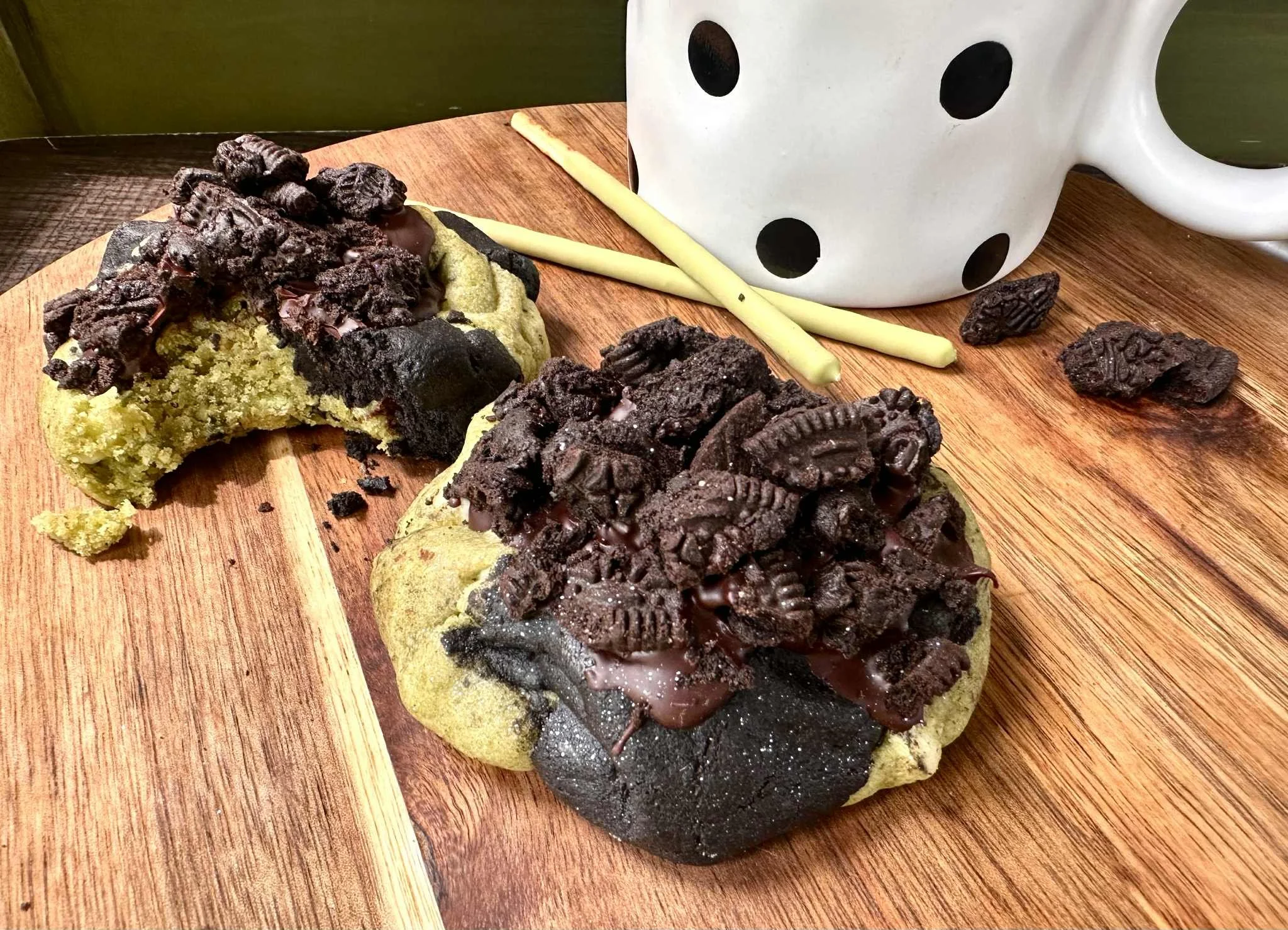 Chocolate chip cookies with Oreo cookies broken and placed on top, a white mug with black polka dots, and white chocolate sticks on a wooden surface.