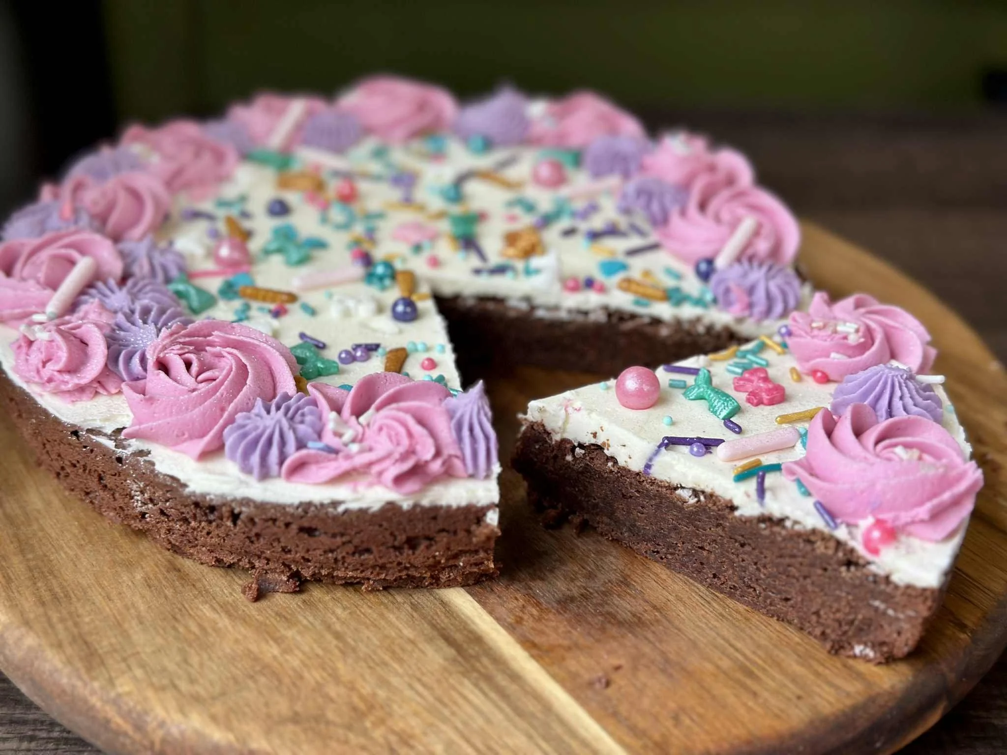 Close-up of a half-eaten chocolate cake with white frosting, decorated with pink and purple frosting roses and colorful sprinkles on a wooden surface.