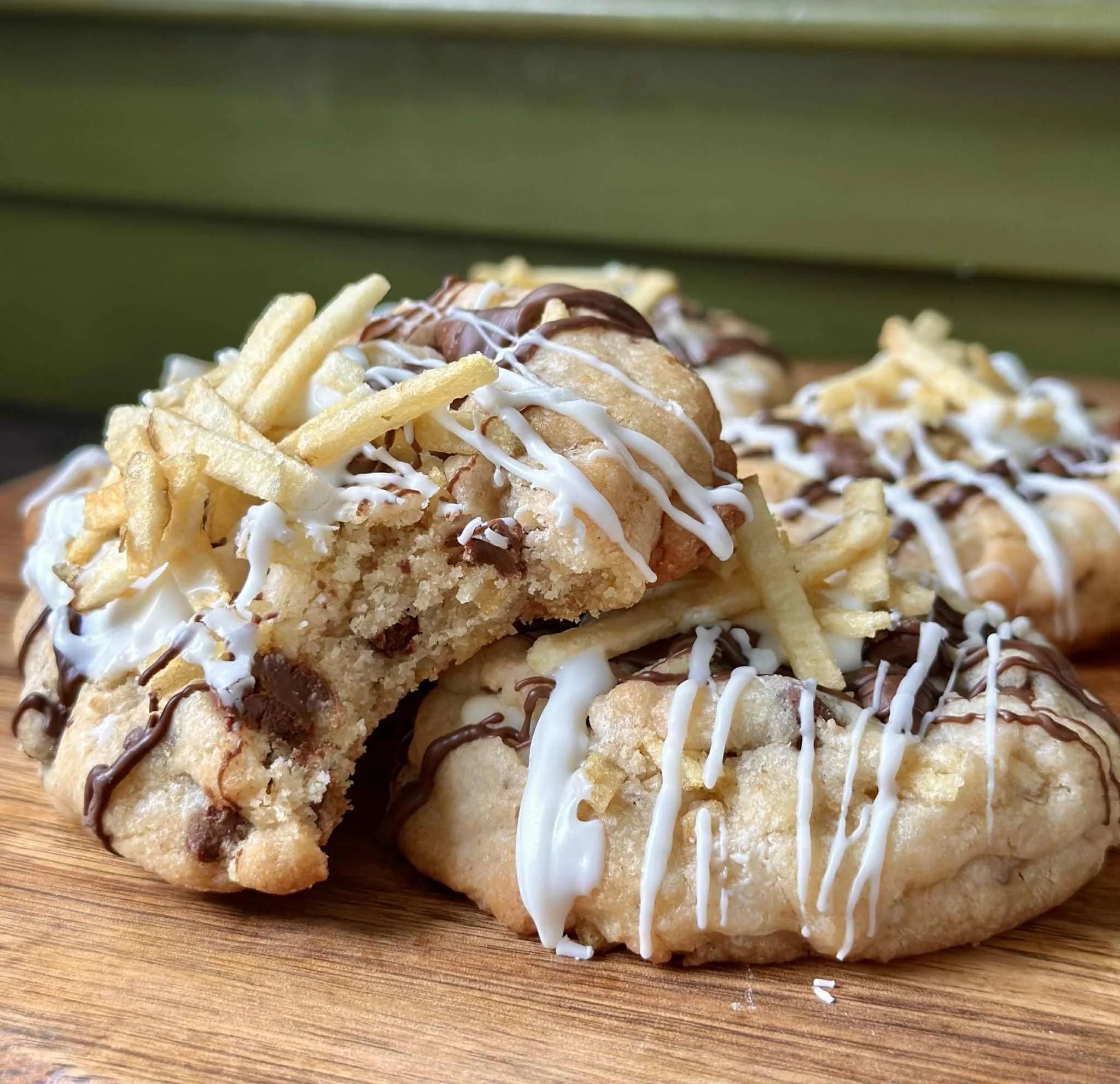 Close-up of chocolate chip cookies topped with white and dark chocolate drizzle and crispy French fries, arranged on a wooden surface.