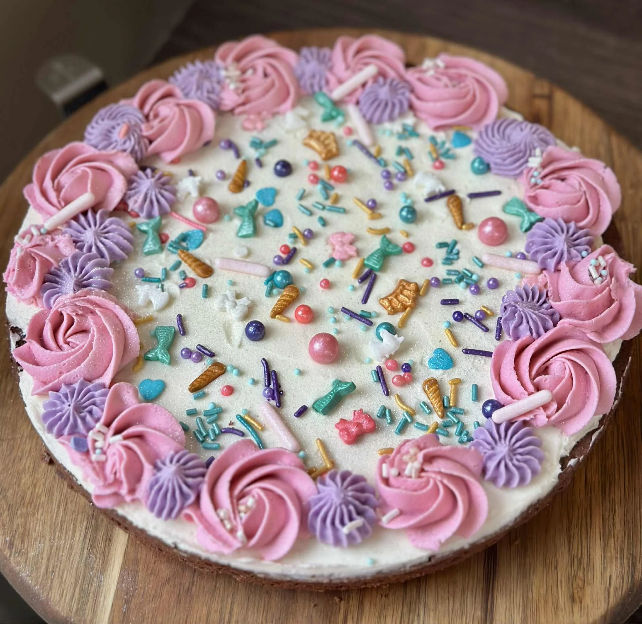 A round birthday cake decorated with pink and purple frosting roses around the edge, topped with colorful sprinkles, candies, and edible decorations on white frosting.