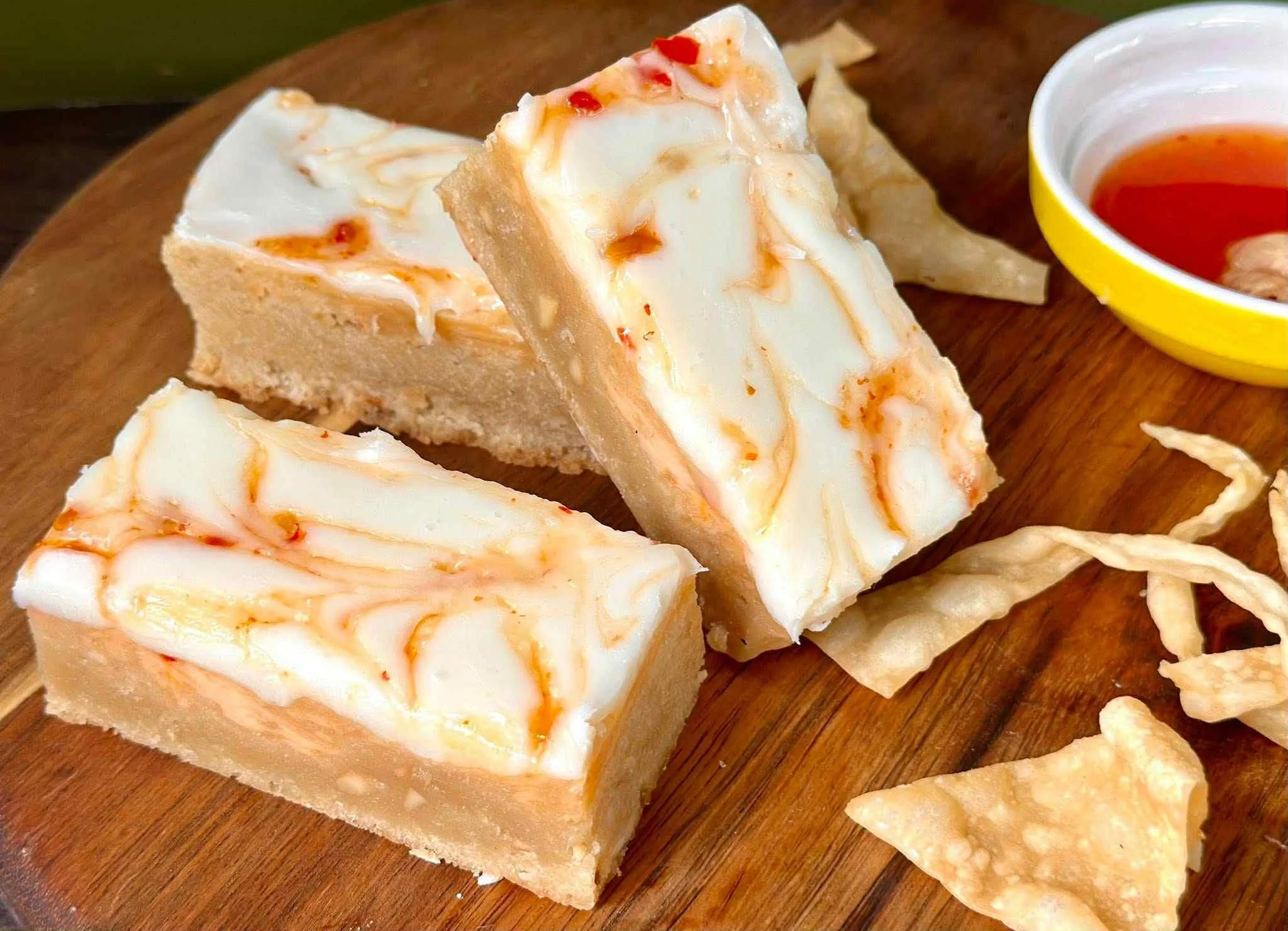 Slices of white cheese with a caramel drizzle on top, served on a wooden platter with a small bowl of red sauce, crispy chips, and tortilla chips.